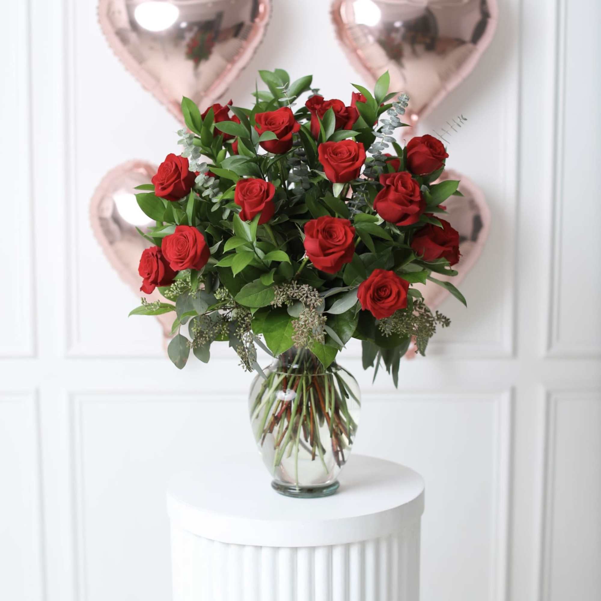 Red roses in a glass vase with pink heart balloons behind them