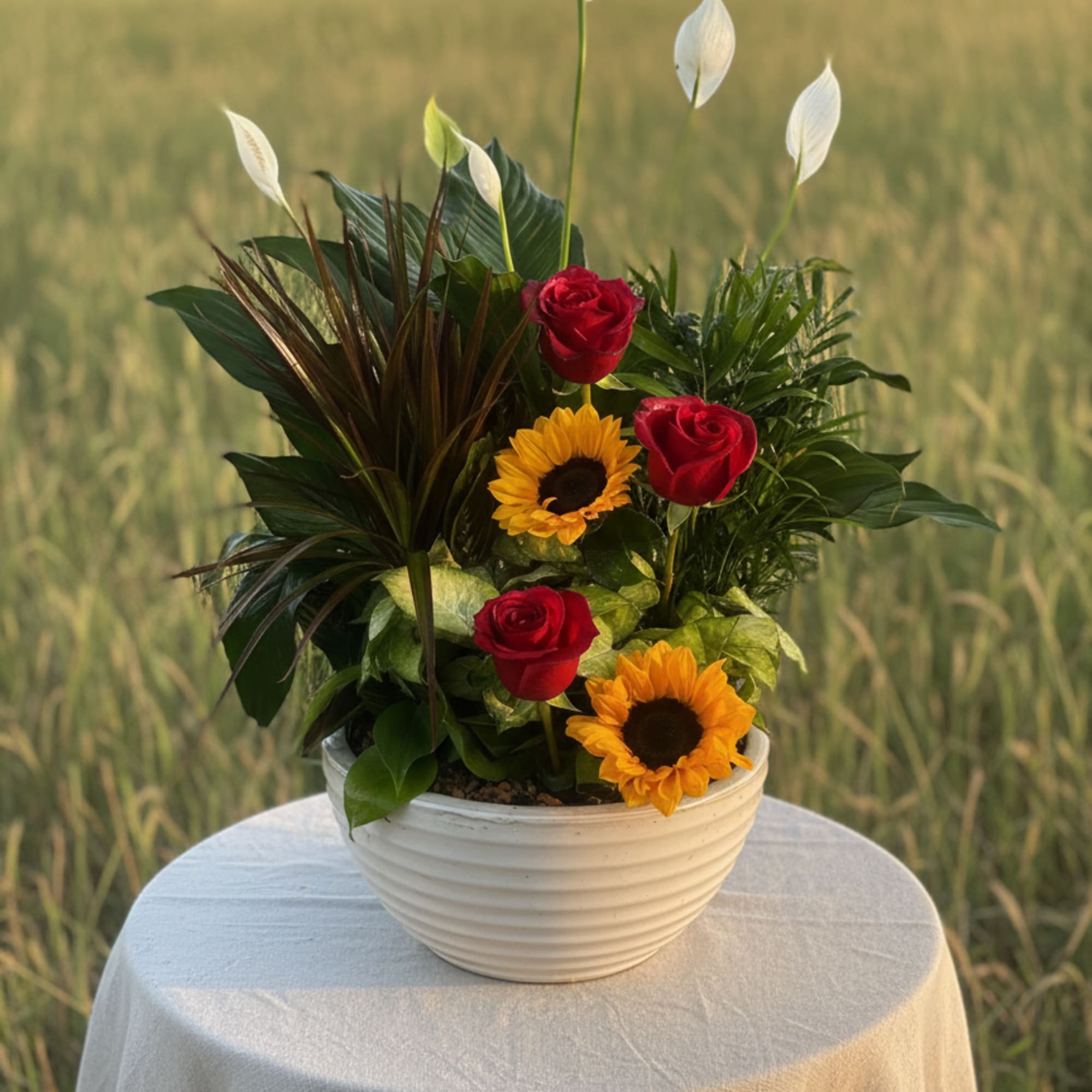 Mixed planter with red roses, yellow sunflowers, and white blooms in a white bowl on a draped table outdoors