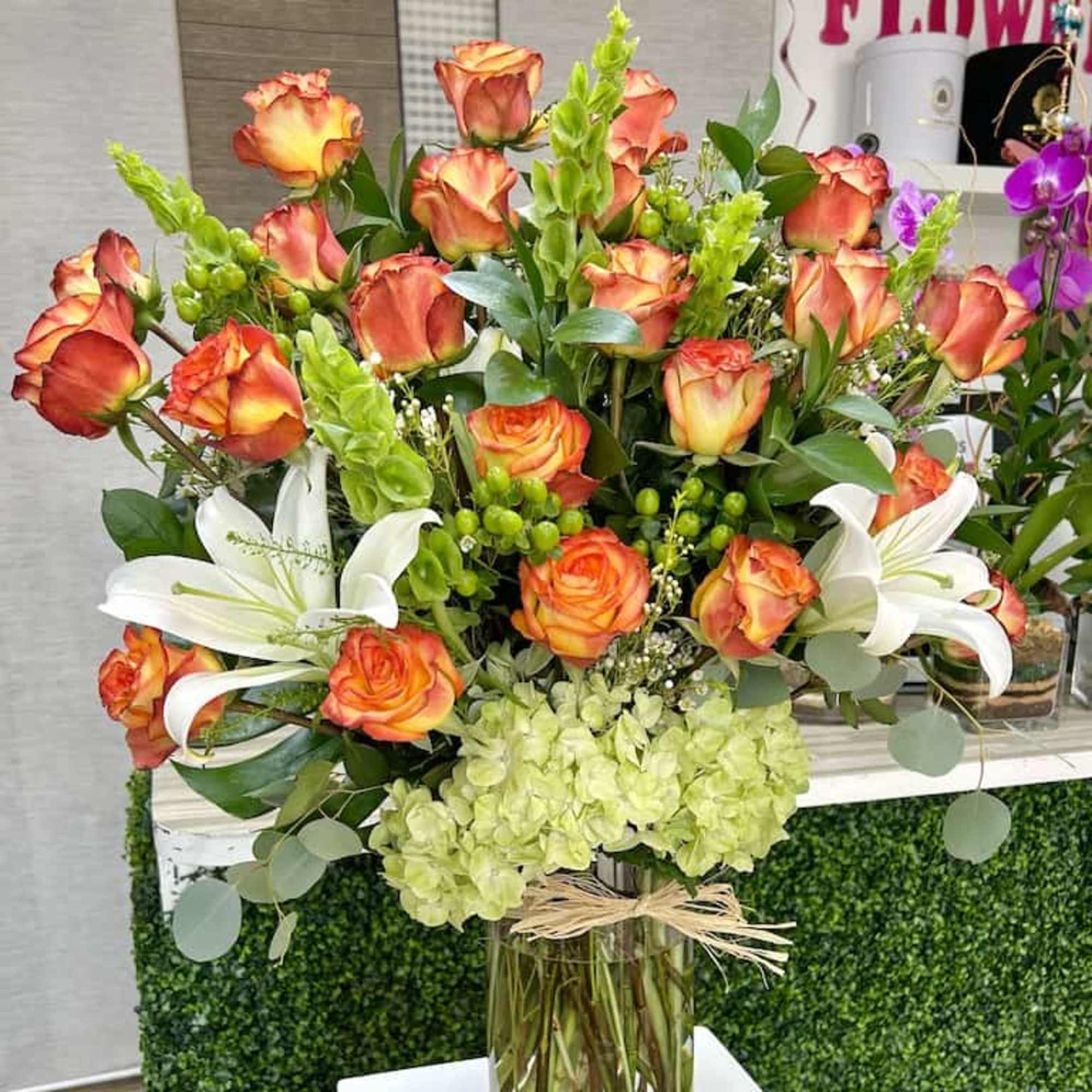 Tall arrangement of orange roses, white lilies, and green hydrangeas in a clear glass vase