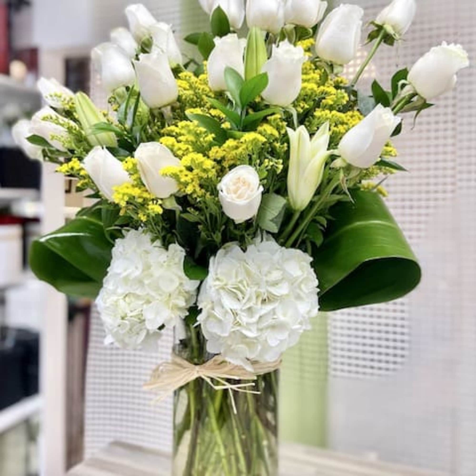 Tall arrangement of white roses, lilies, and hydrangeas with yellow flowers and large green leaves in a glass vase.