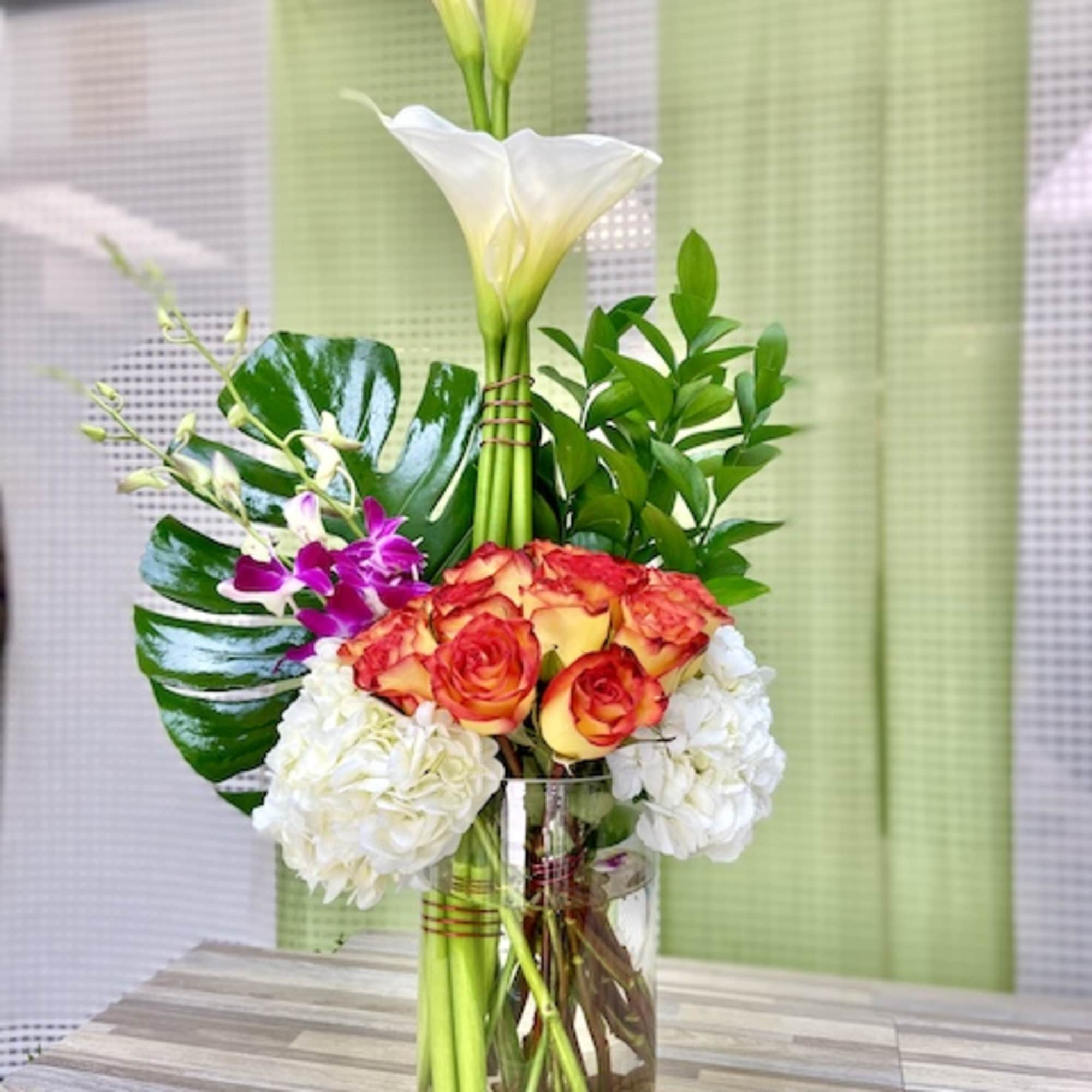 Tall arrangement of white calla lilies, orange roses, hydrangeas, and orchids in a glass vase with stones.