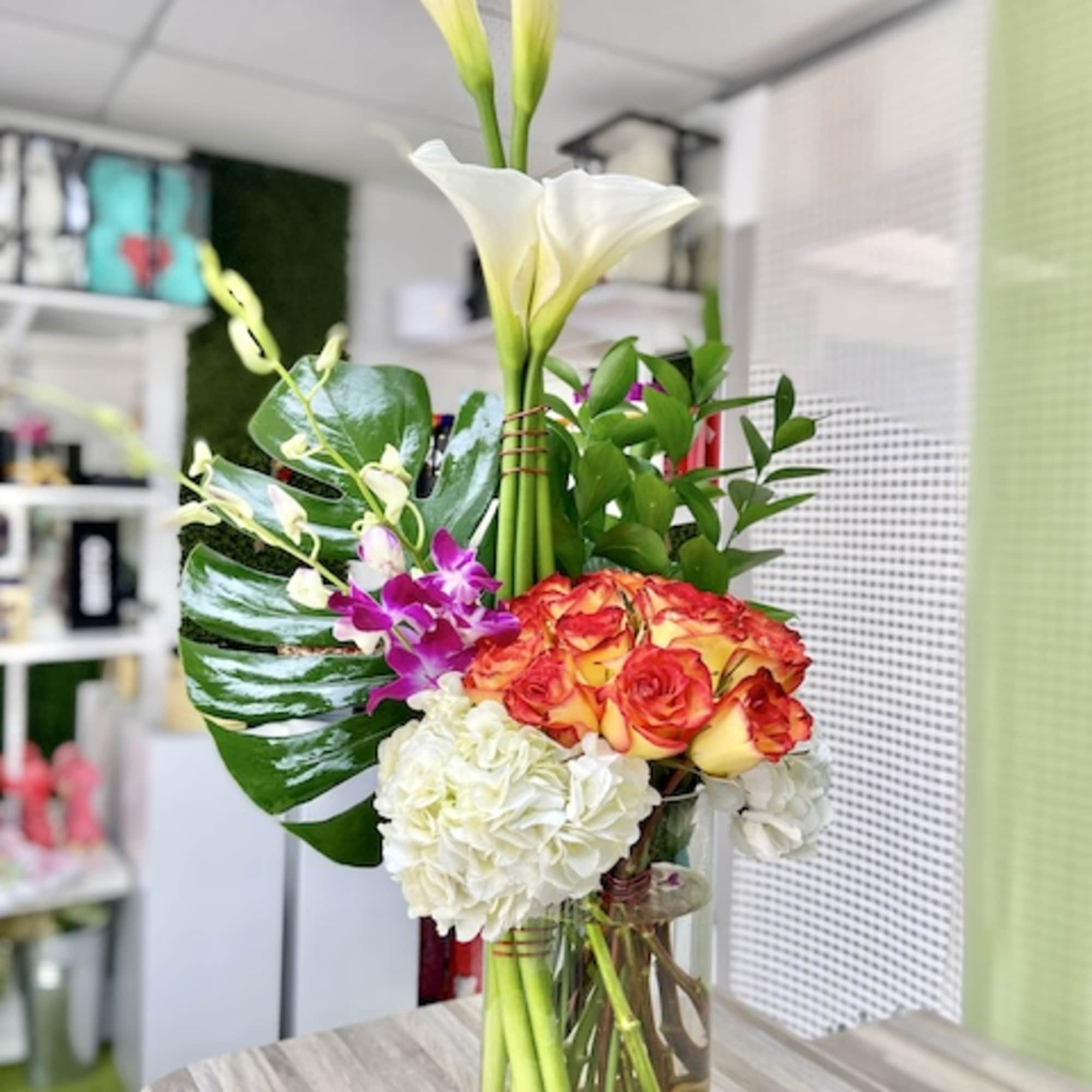 Tall arrangement of white calla lilies, orange roses, orchids, and hydrangea in a glass vase with stones