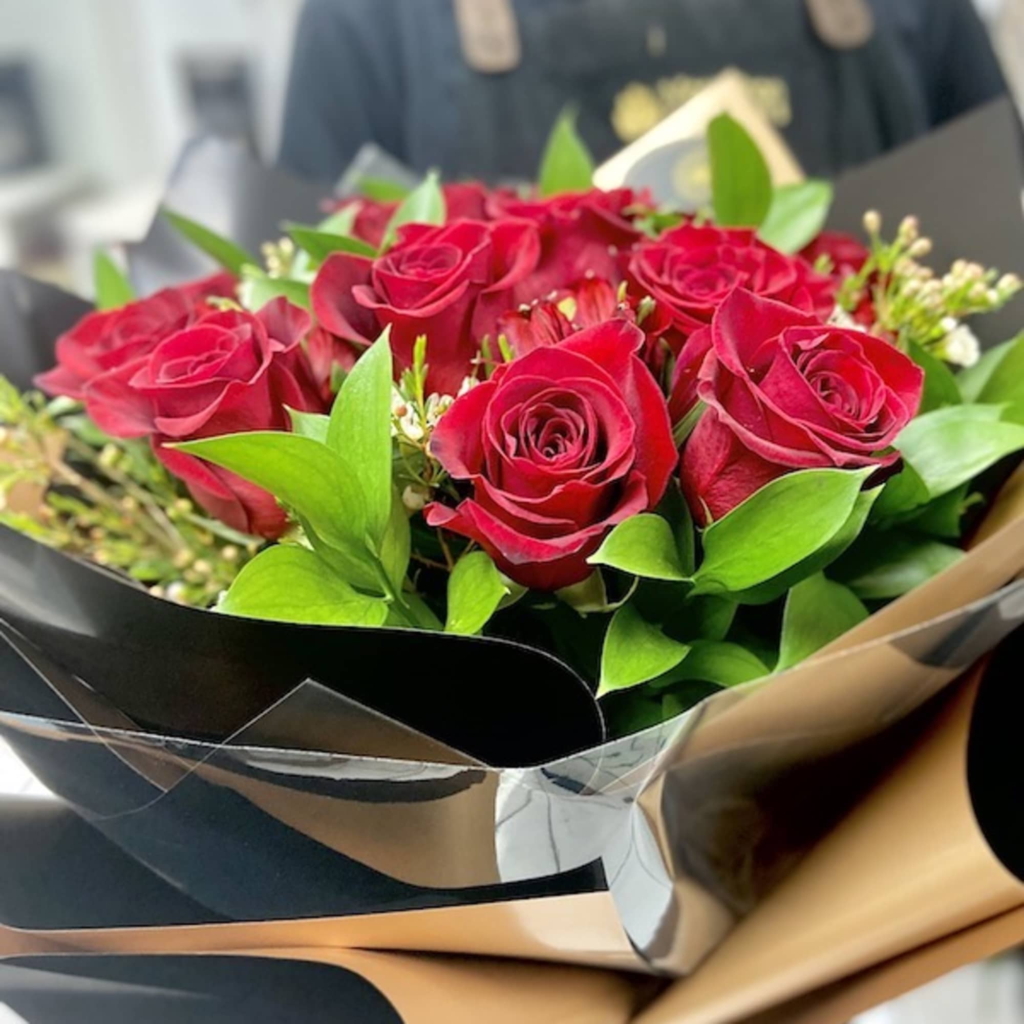 Hand-tied bouquet of red roses with filler flowers wrapped in black and tan paper.
