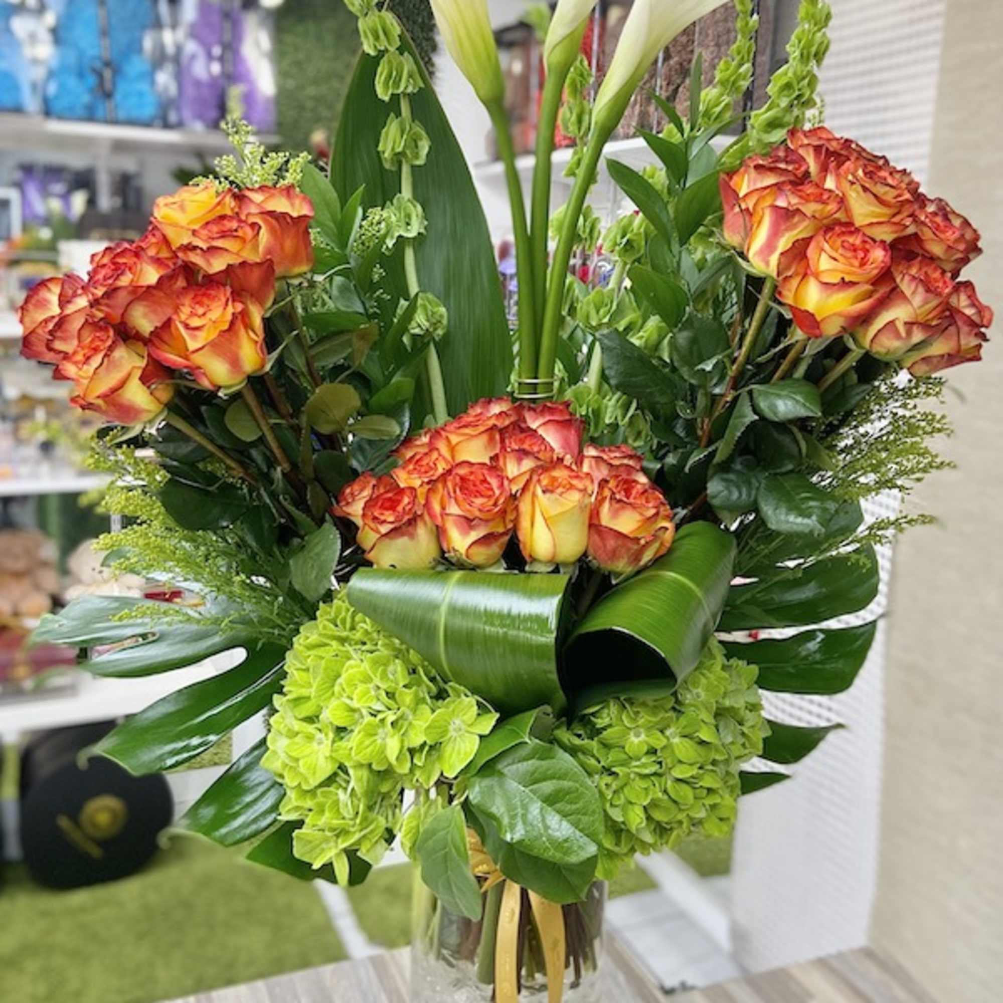 Tall arrangement of orange-yellow roses, white calla lilies, and green hydrangeas in a clear glass vase