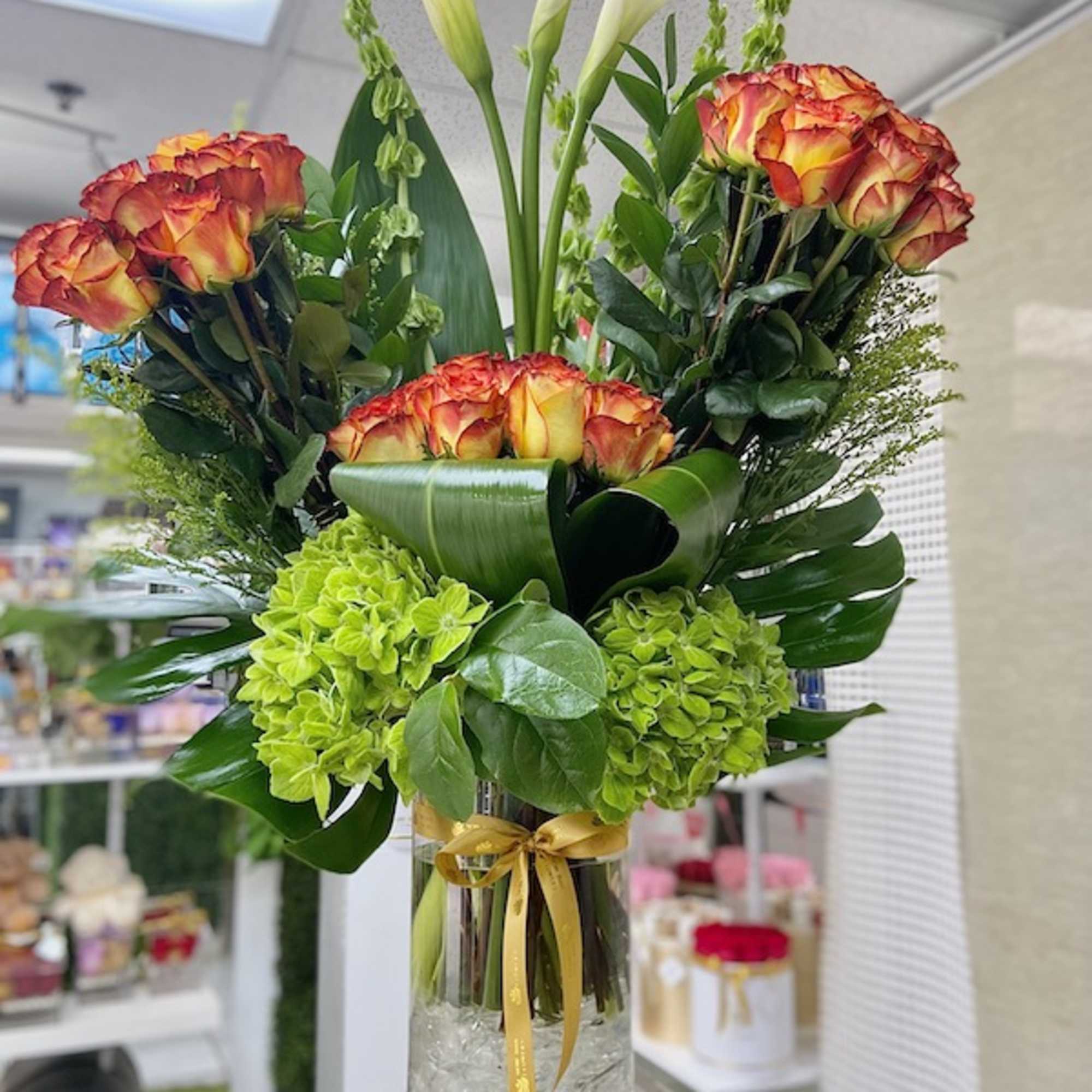 Tall arrangement of orange roses, white calla lilies, and green hydrangeas in a clear vase with gold ribbon