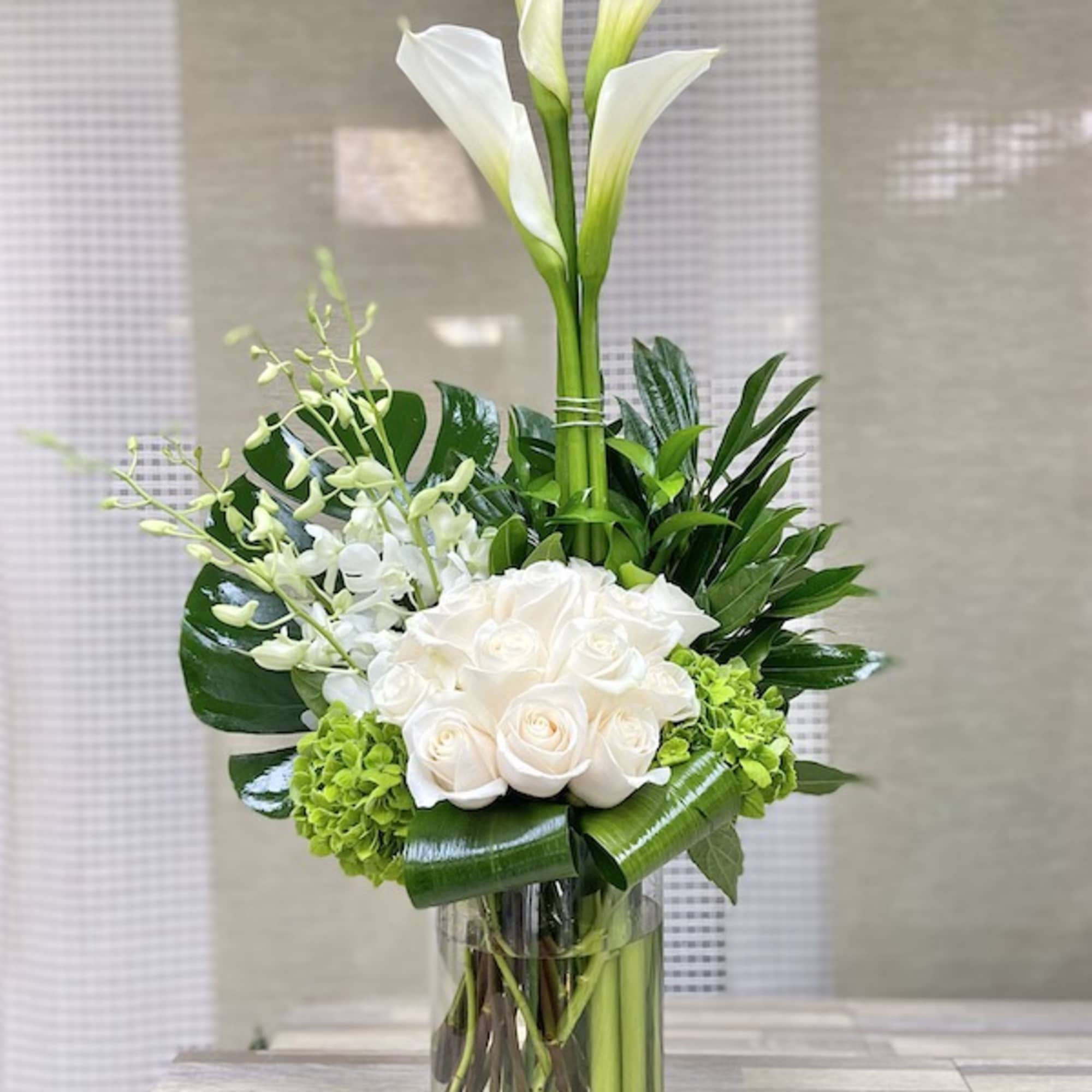 Tall white calla lilies, roses, orchids, and green accents arranged in a clear glass cylinder vase.