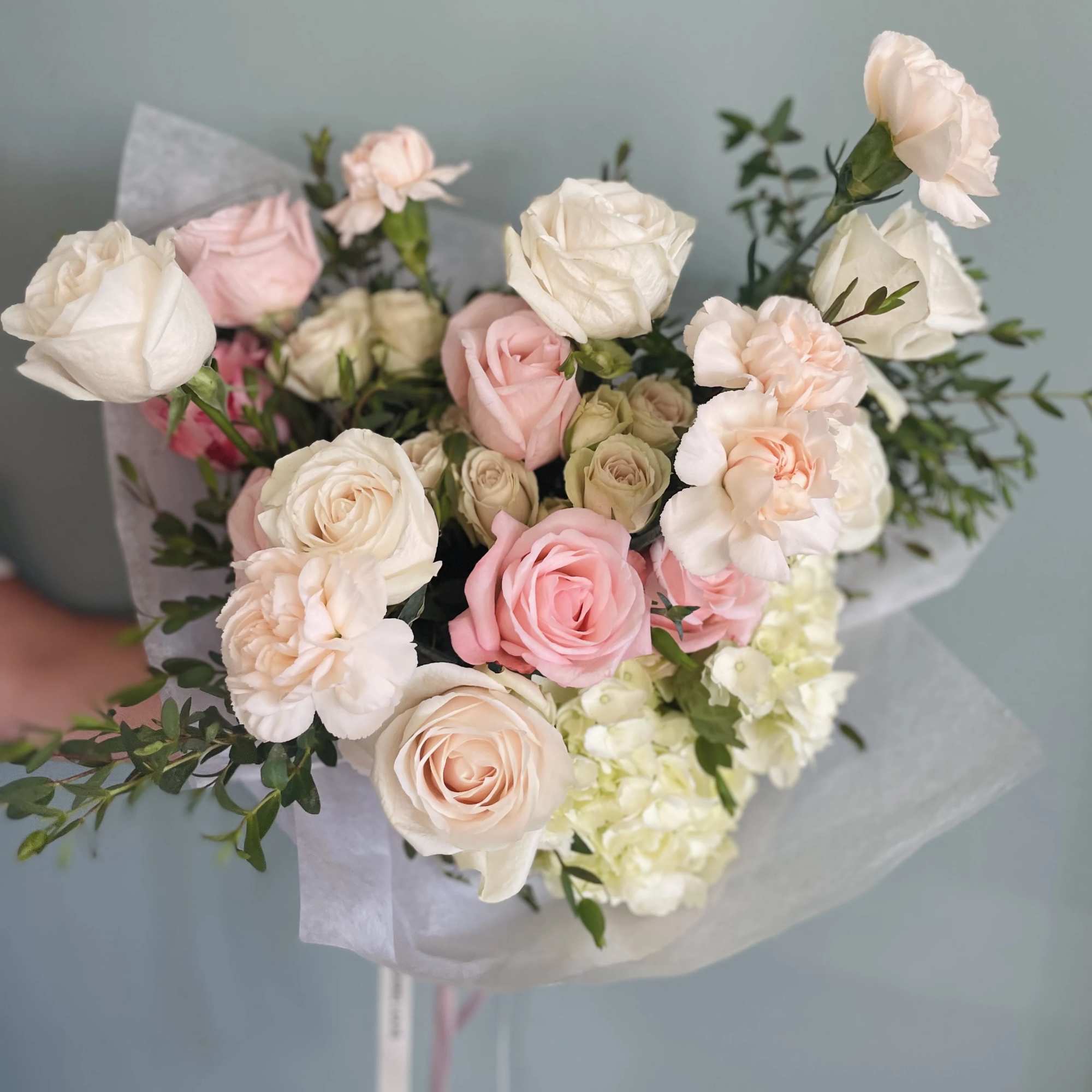 Hand-tied bouquet of white and blush roses with soft pink carnations wrapped in white tissue paper.