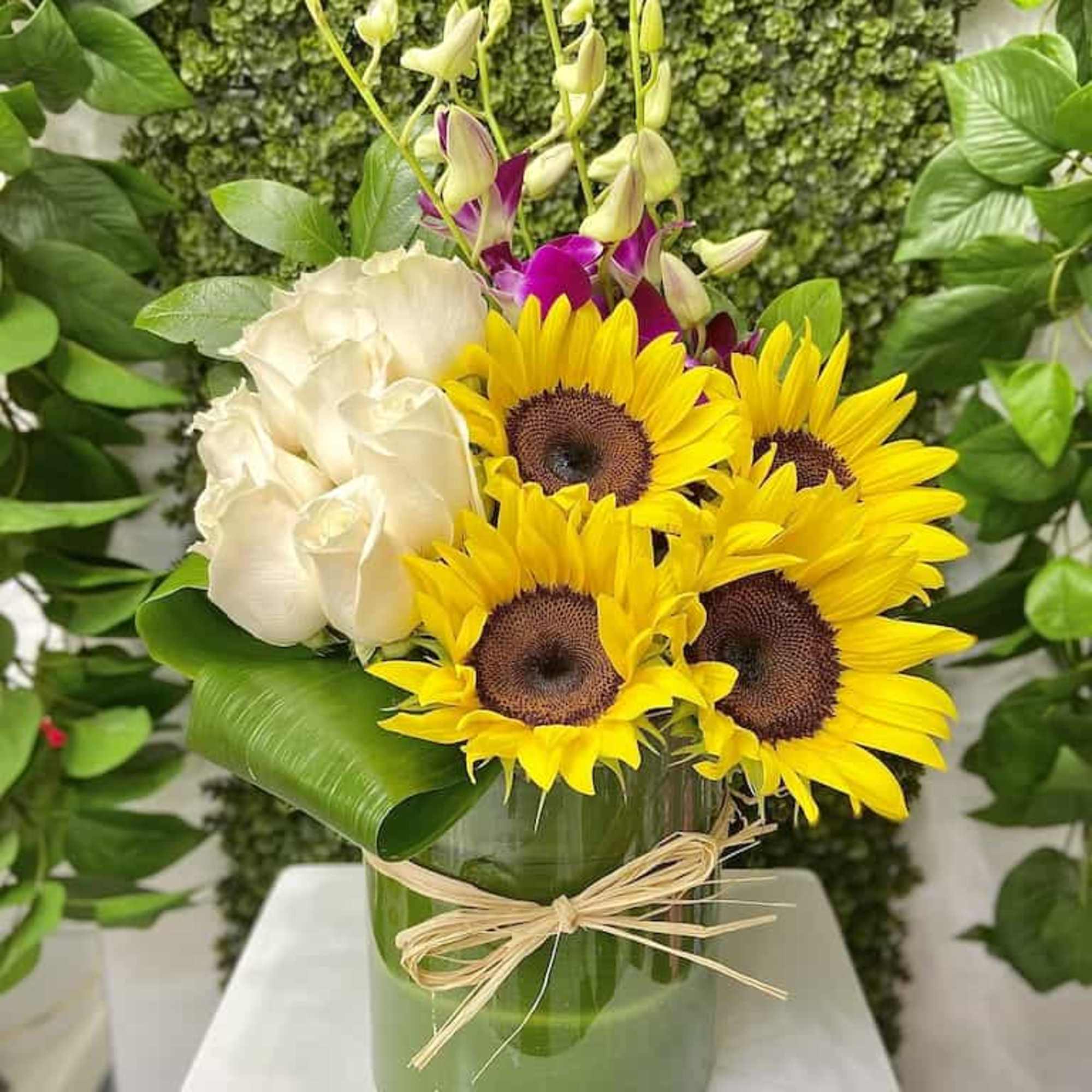 Sunflowers, white roses, and purple orchids in a glass vase with a large green leaf wrap.