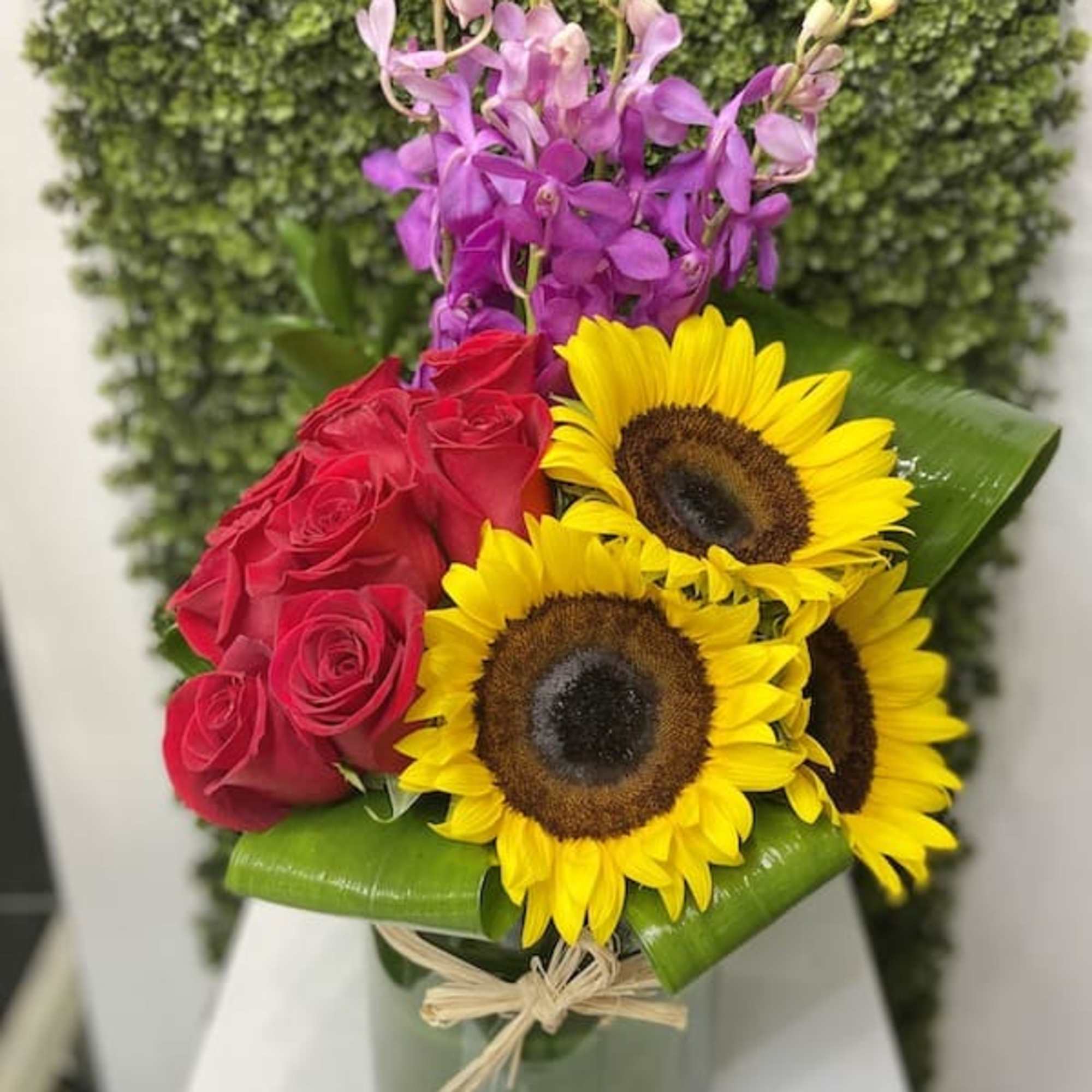 Arrangement of yellow sunflowers, red roses, and purple orchids in a glass vase with leaf accents