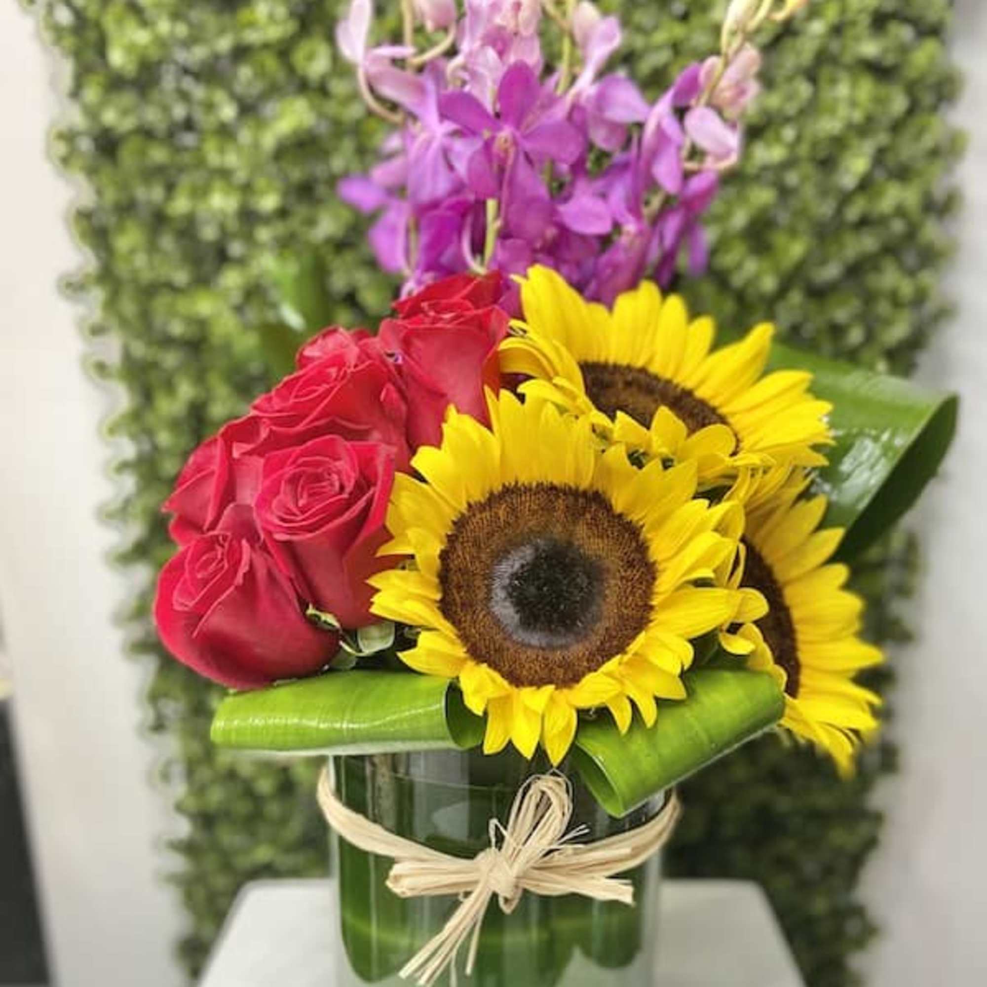 Arrangement of sunflowers, red roses, and purple orchids in a clear glass vase with a raffia bow.