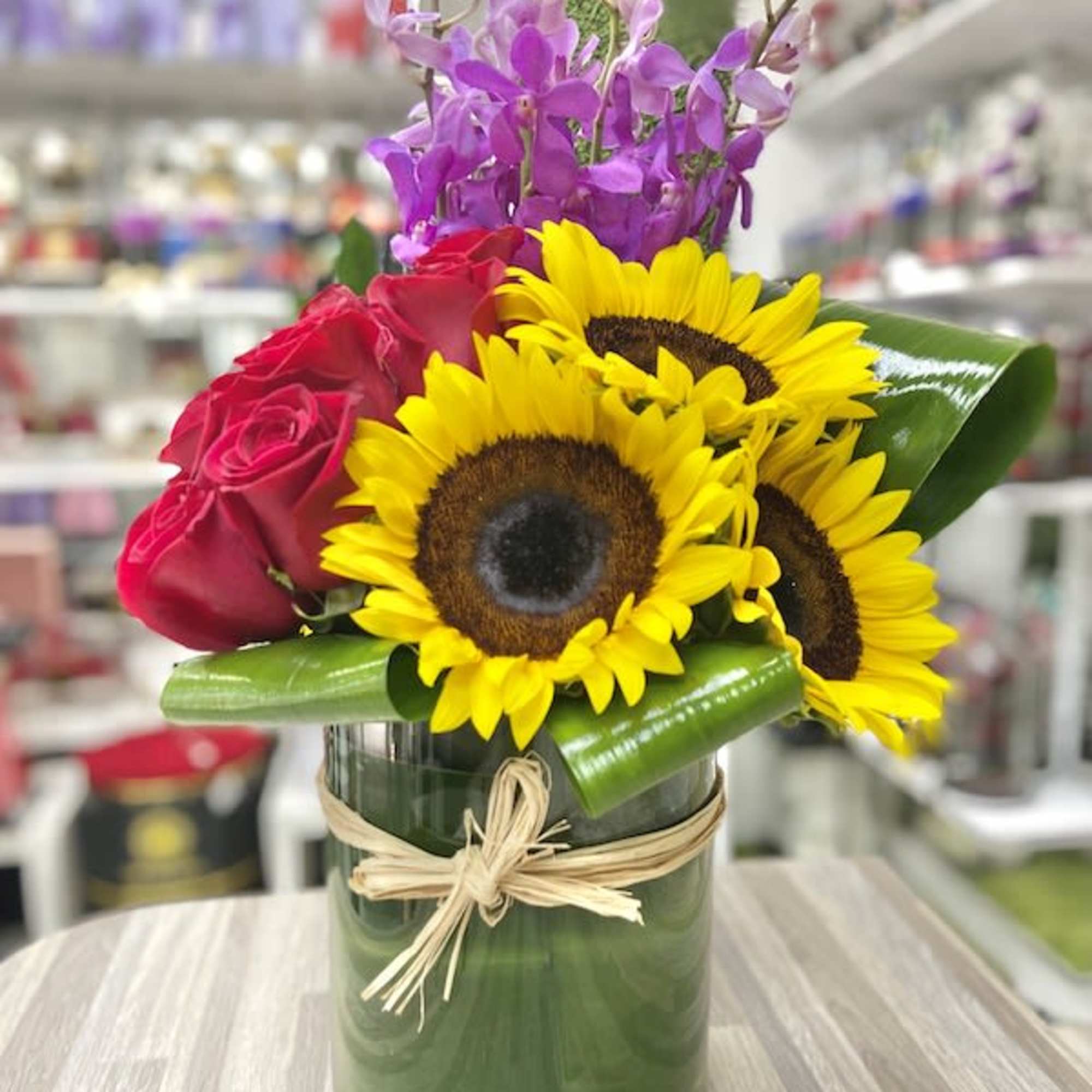 Mixed arrangement of sunflowers, red roses, and purple orchids in a glass cylinder vase with raffia bow