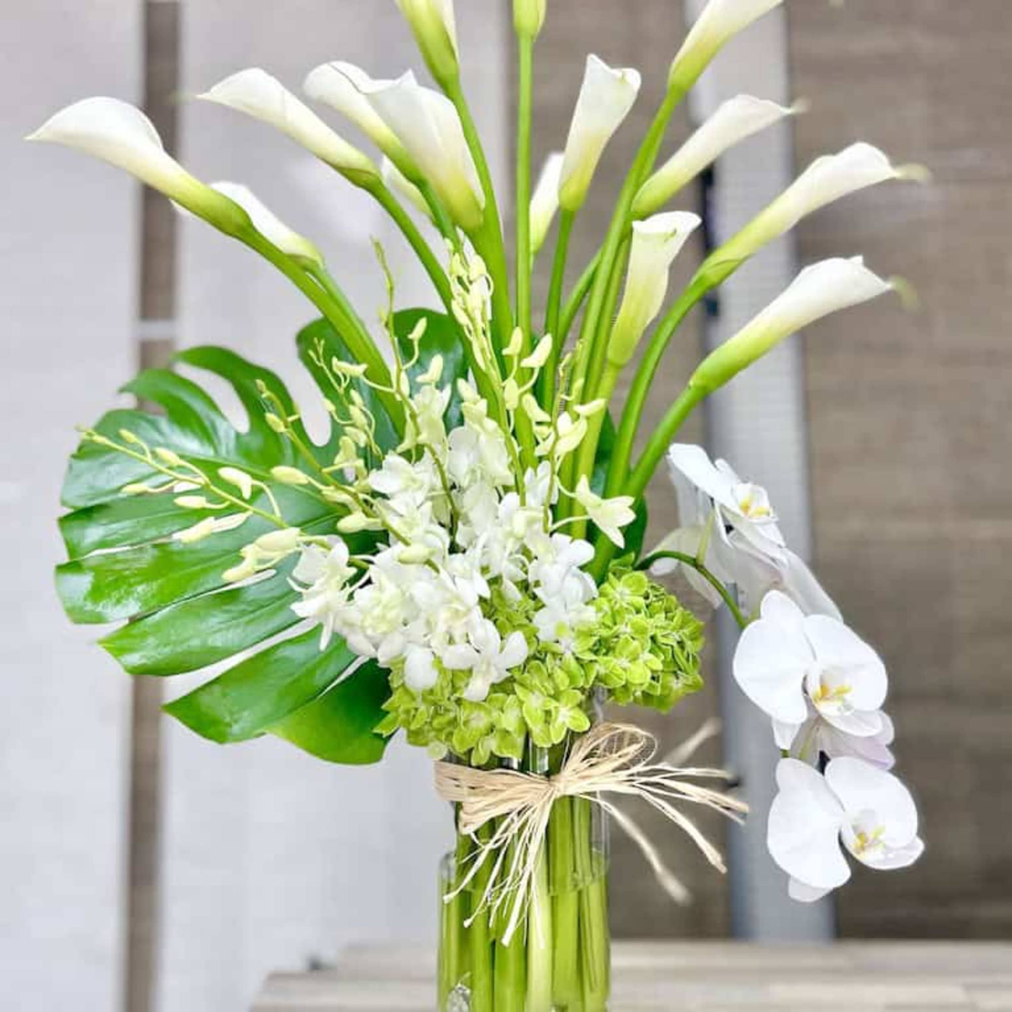 Tall white calla and orchid arrangement with green hydrangea in a clear glass vase