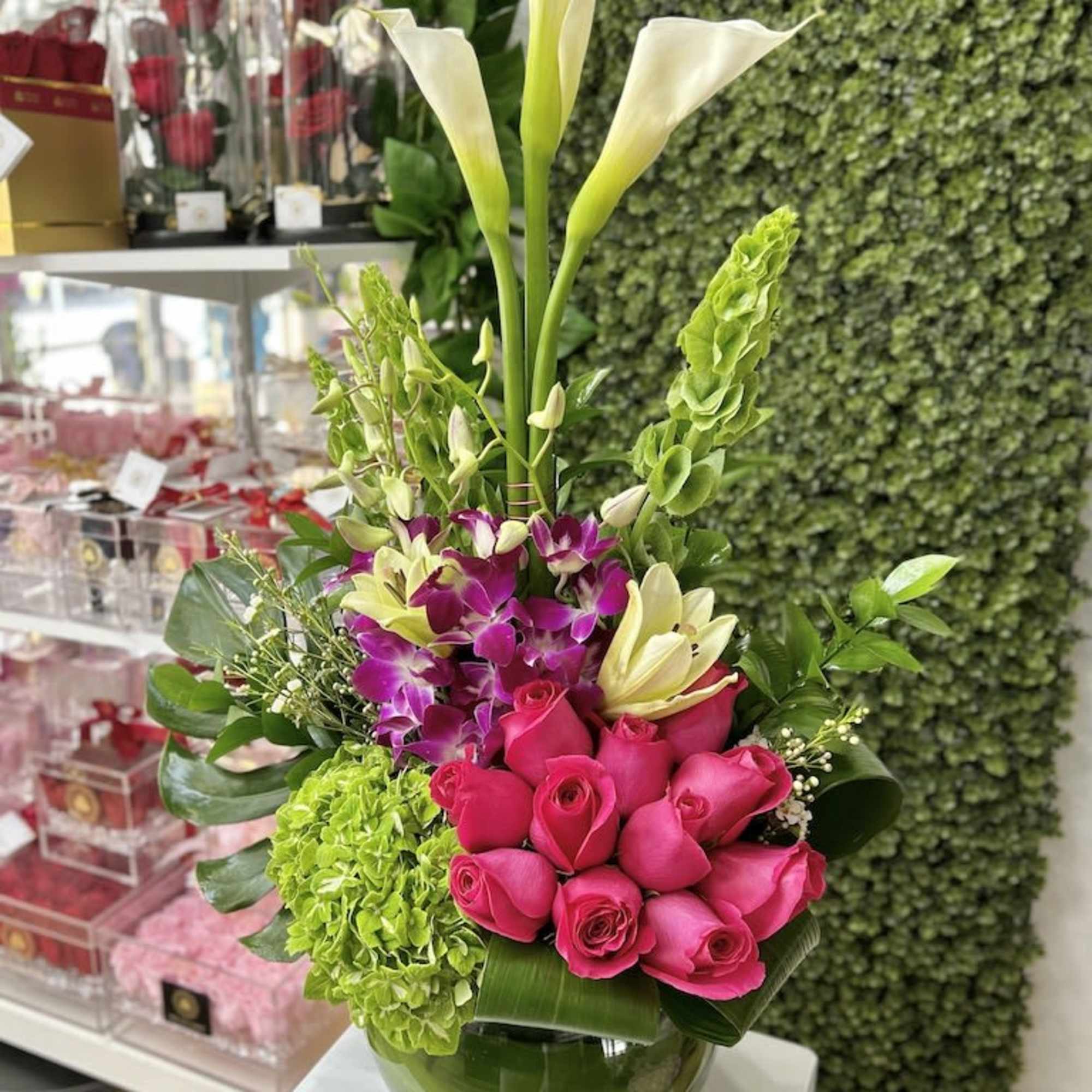 Tall mixed arrangement with white calla lilies, pink roses, orchids and green hydrangea in a round glass vase.