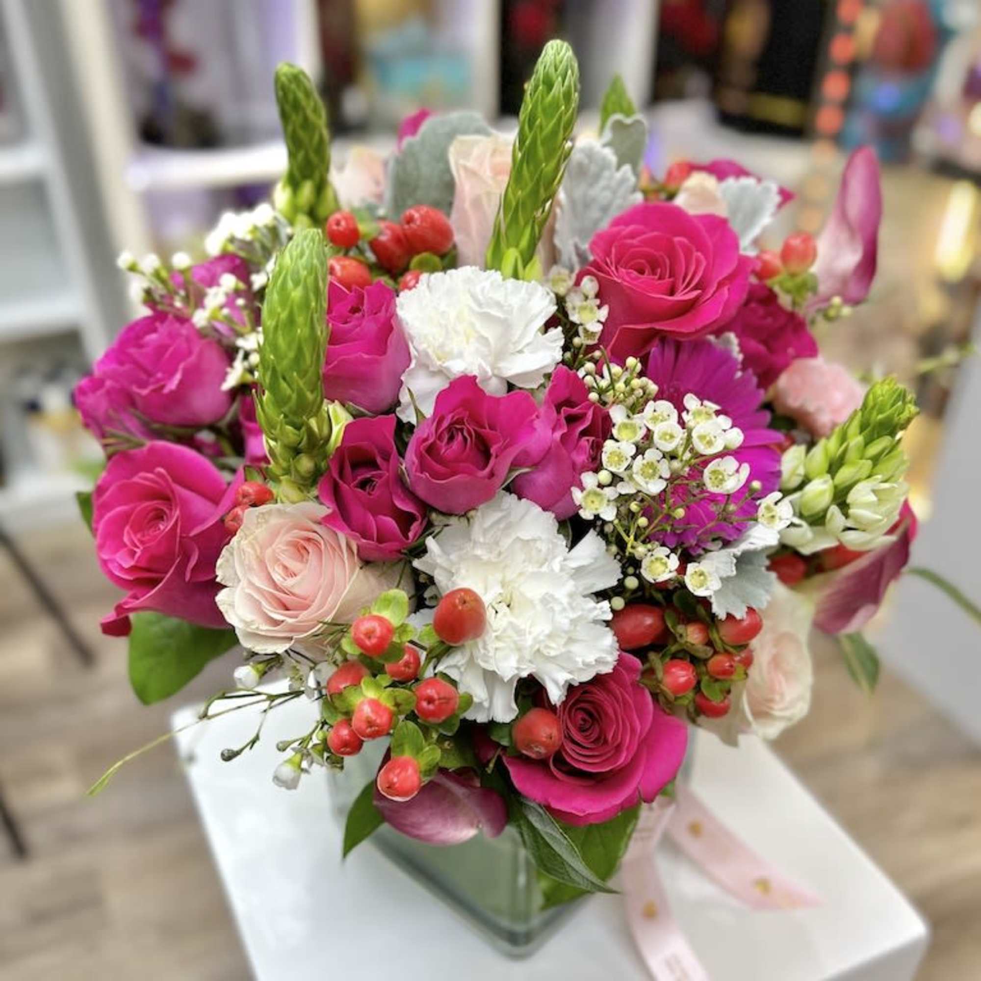 Compact arrangement of hot pink and blush roses with white carnations in a clear glass cube vase.