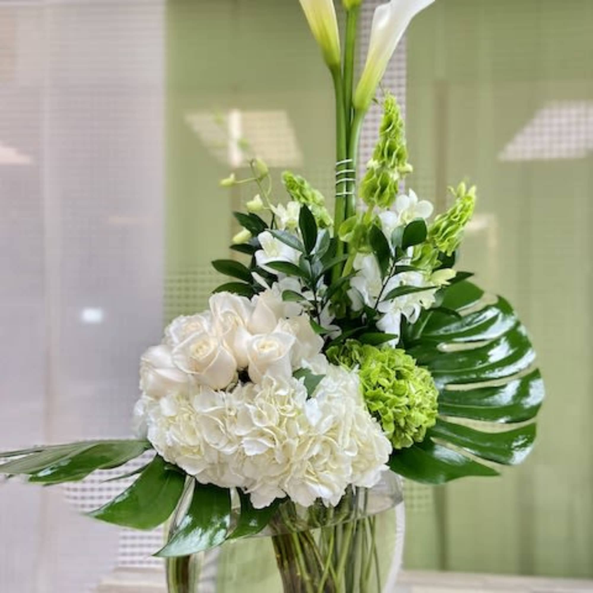 Tall white calla lilies with white roses and hydrangeas in a clear glass vase.