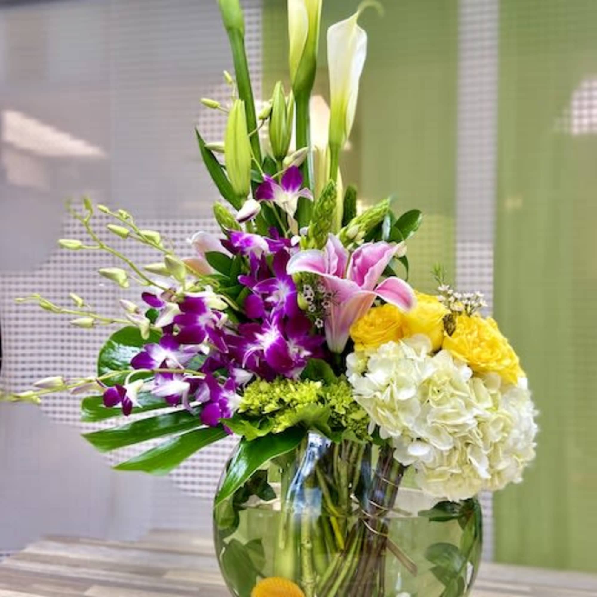 Tall arrangement with white calla lilies, purple orchids, pink lilies and yellow blooms in a round glass vase.