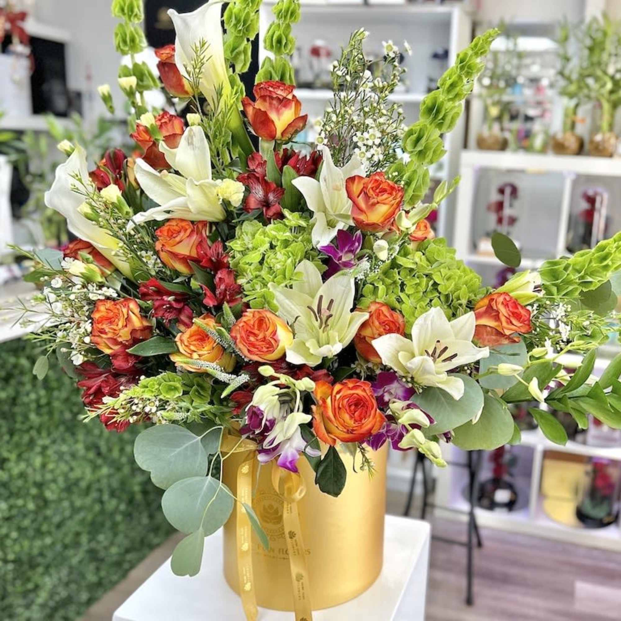 Tall arrangement of orange roses and white lilies in a gold hatbox-style container.