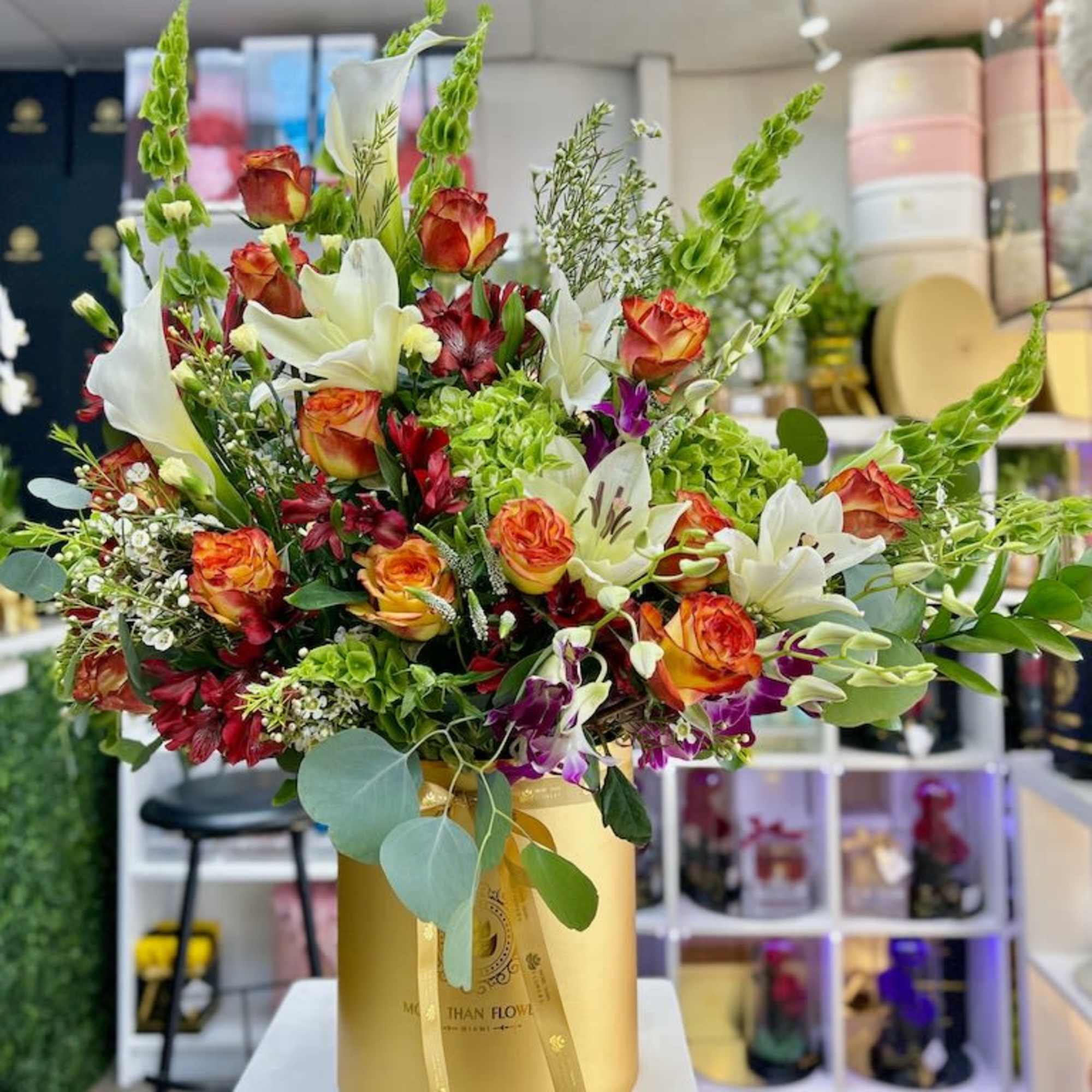 Tall arrangement of orange roses, white lilies, and mixed blooms in a round gold hatbox container