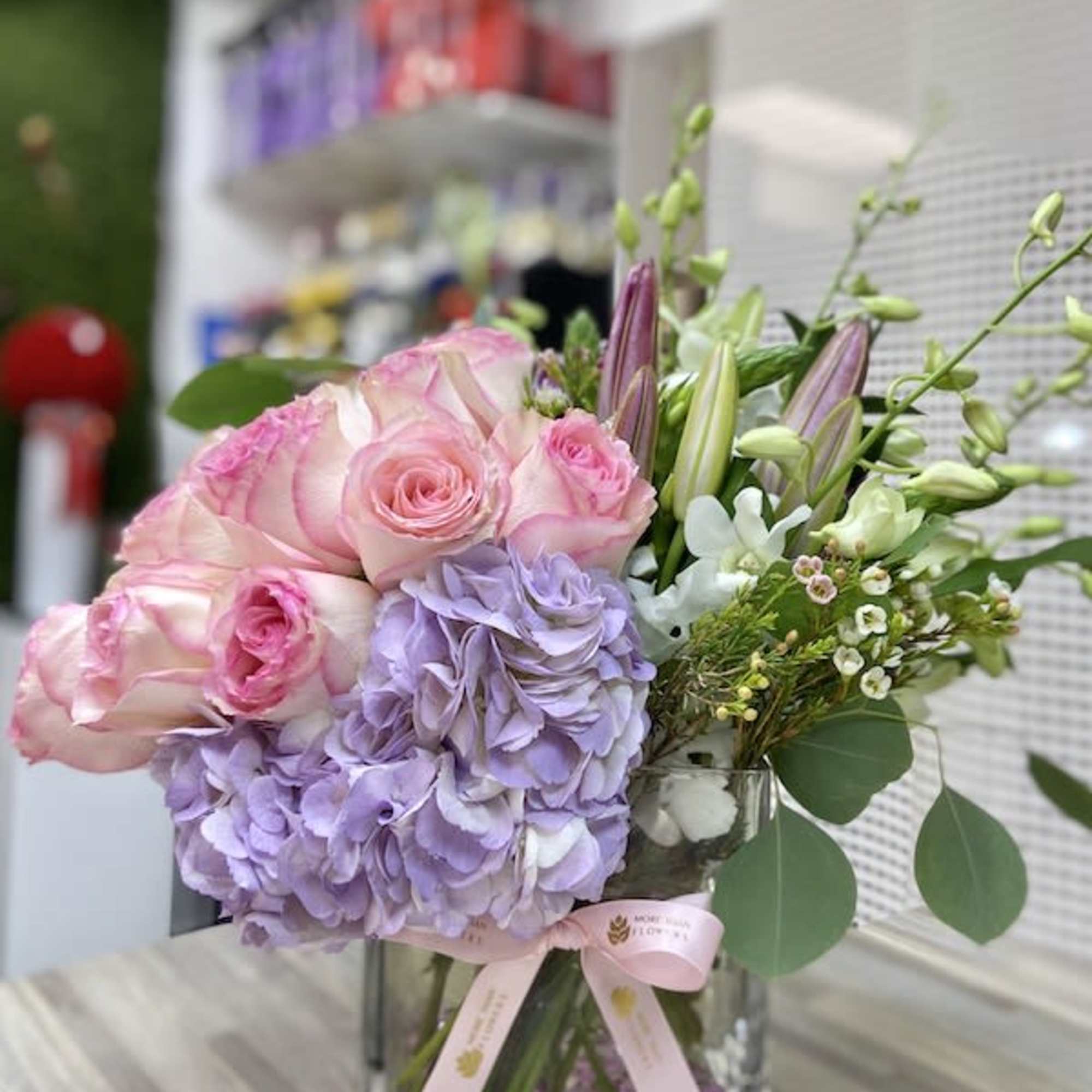 Square glass vase arrangement with pink roses, lavender hydrangeas, and white lilies accented by small white blossoms.