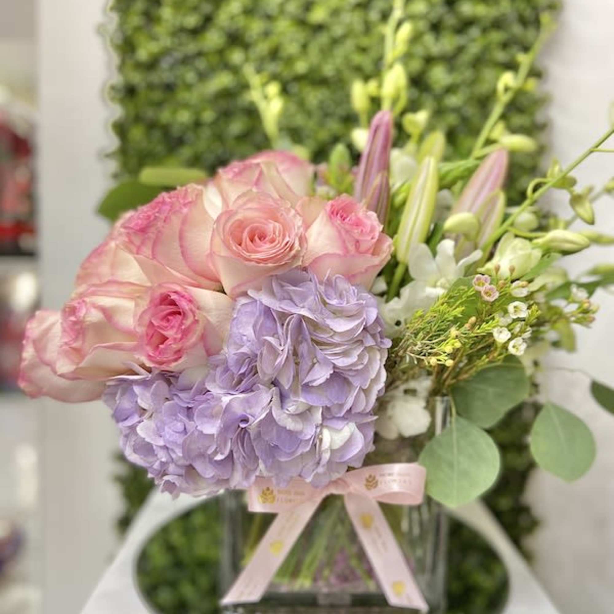Low pastel arrangement of pink roses, lavender hydrangea, and lilies in a clear square vase with ribbon.