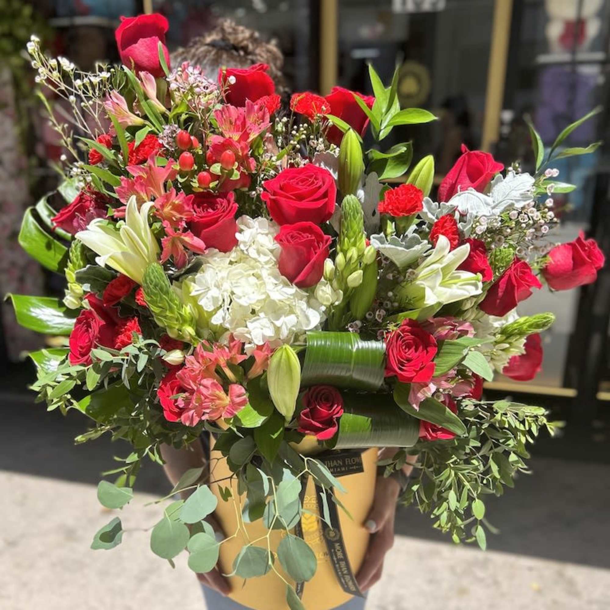 Large arrangement of red roses, lilies, and white hydrangeas in a yellow hatbox-style container