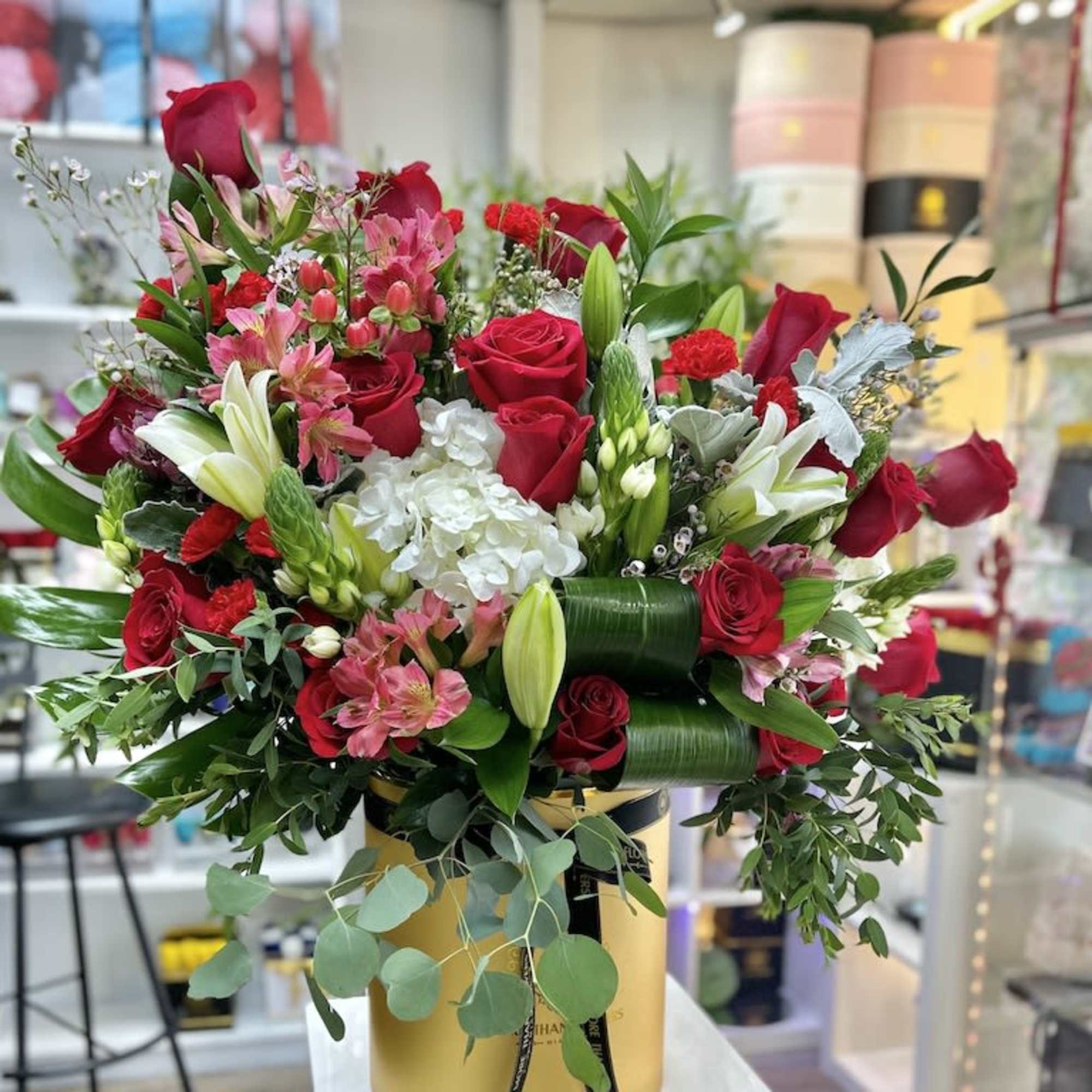 Large arrangement of red roses, lilies, and mixed blooms in a round gold hatbox.