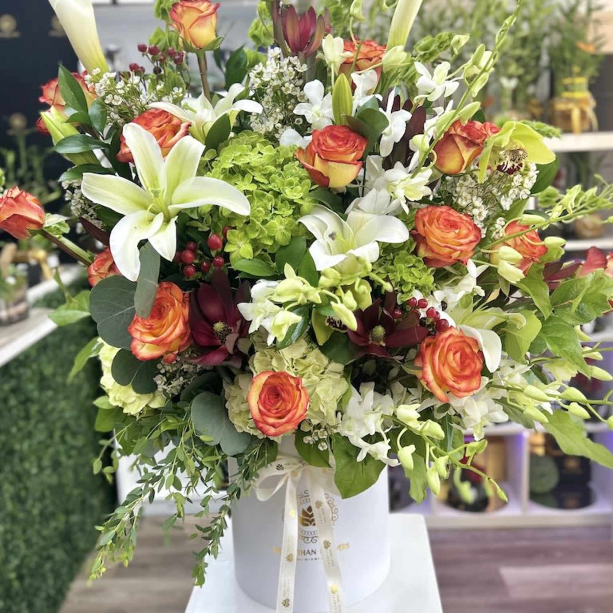 Tall mixed arrangement of orange roses, white lilies, orchids, and green hydrangeas in a white hat box.