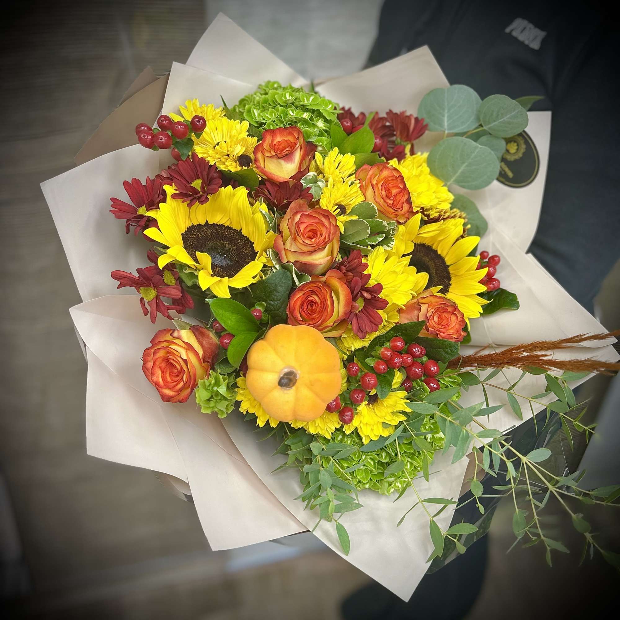 Hand-tied bouquet with sunflowers, orange roses, yellow mums and a small pumpkin accent.