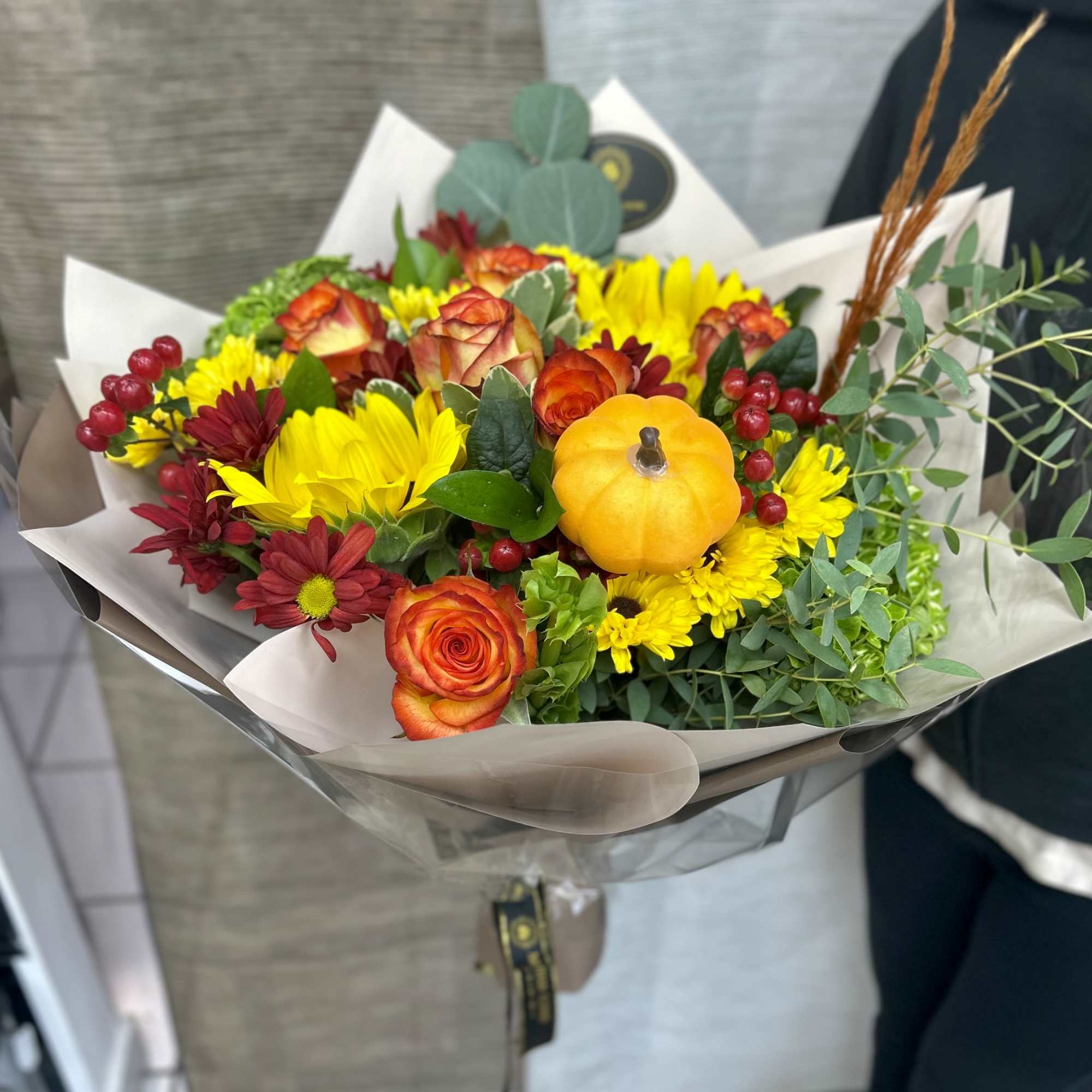 Hand-tied bouquet of yellow mums, orange roses, red flowers and berries with a small decorative pumpkin.