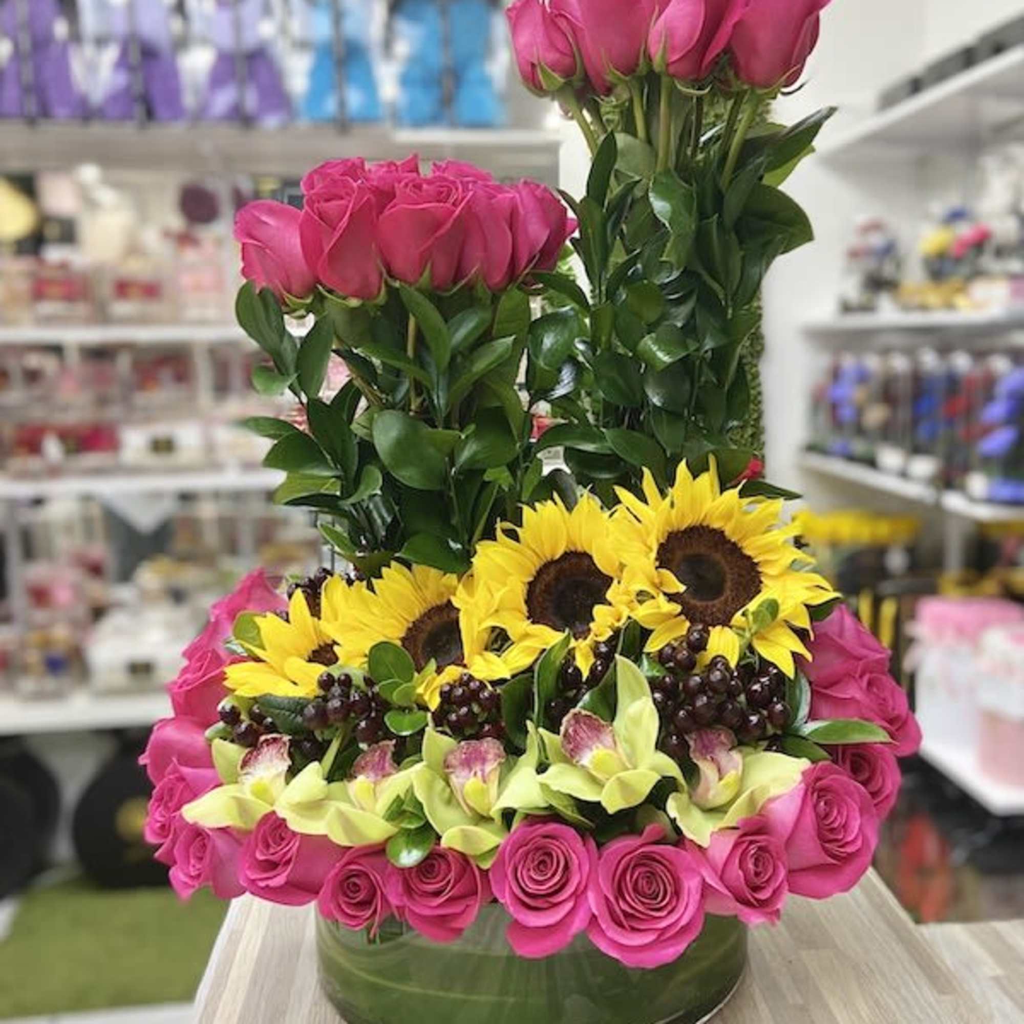 Low glass vase of hot pink roses, yellow sunflowers, green orchids, grapes, and tall pink rose clusters.