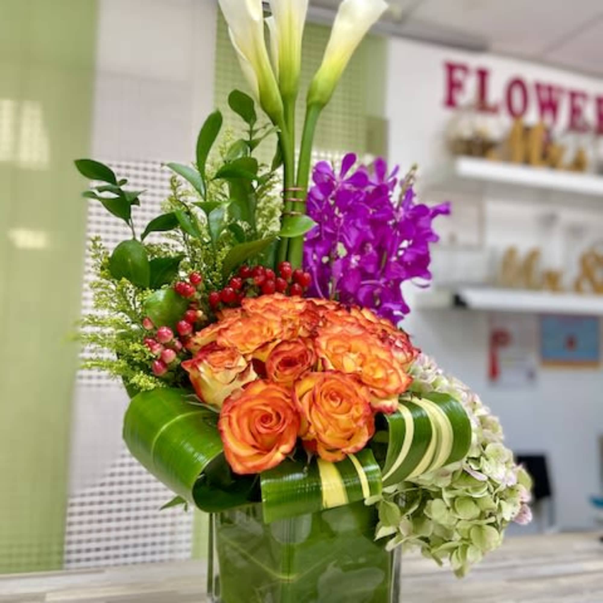 Tall modern arrangement with white calla lilies, orange roses and purple orchids in a green-lined glass cube vase.