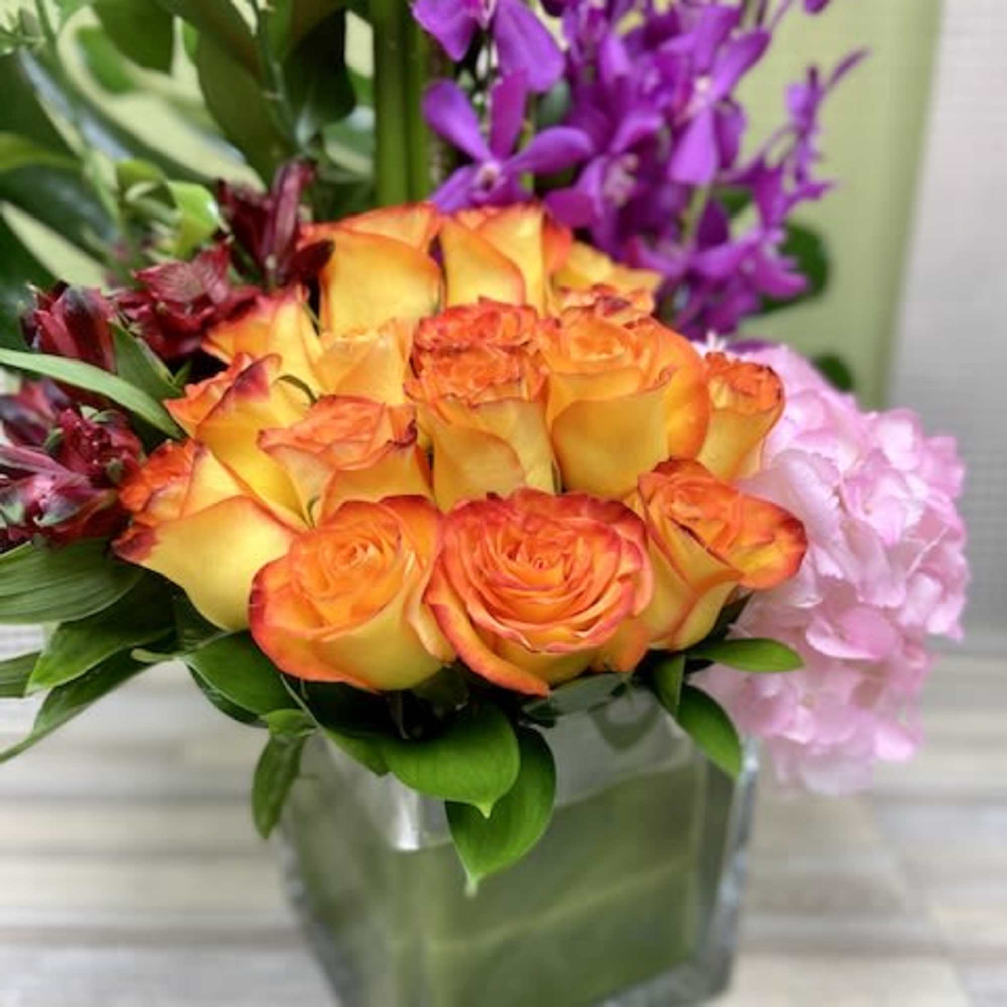 Orange and yellow roses with purple orchids and pink hydrangea in a glass cube vase