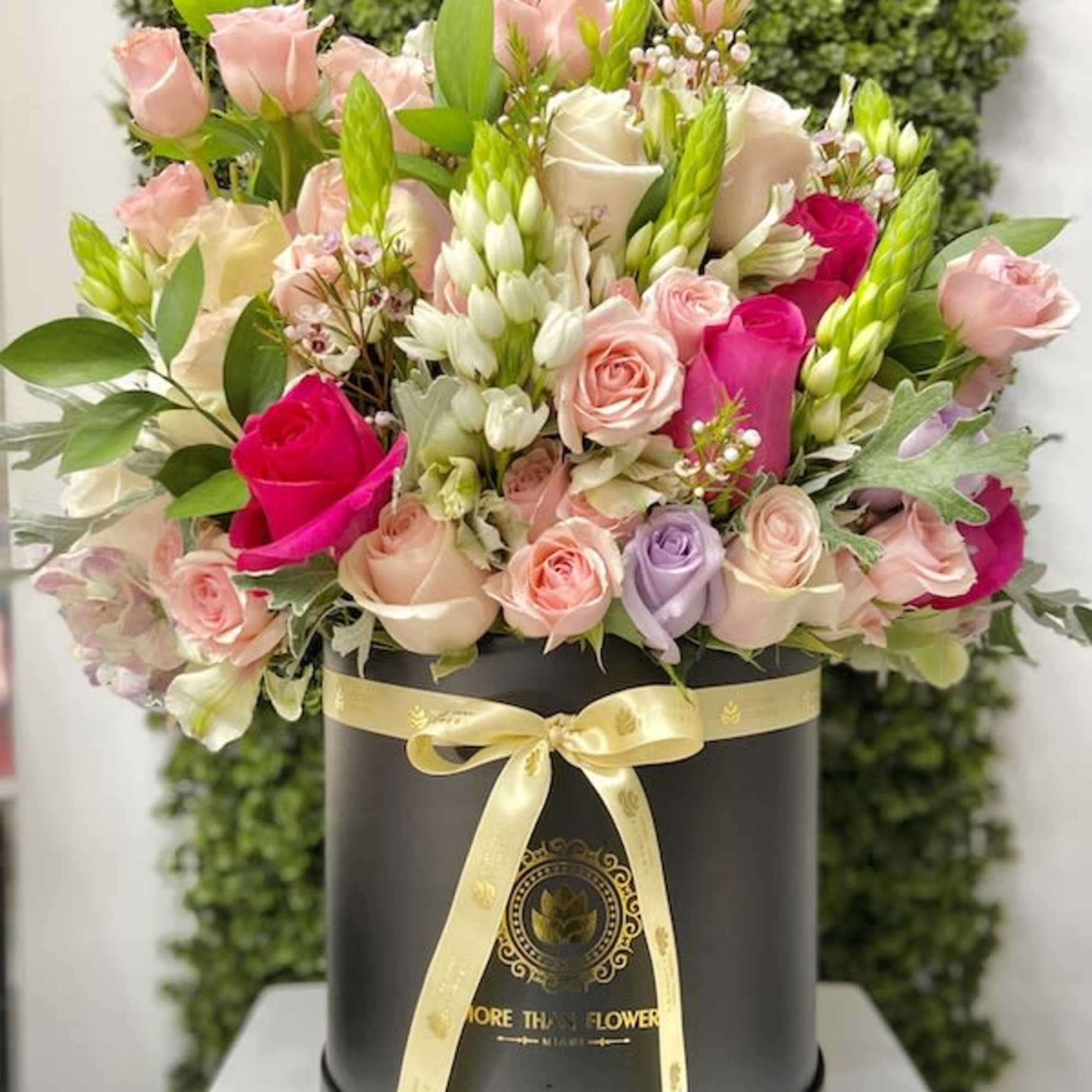 Round hatbox of mixed pink, cream, and purple roses with white accent flowers and a yellow ribbon bow.