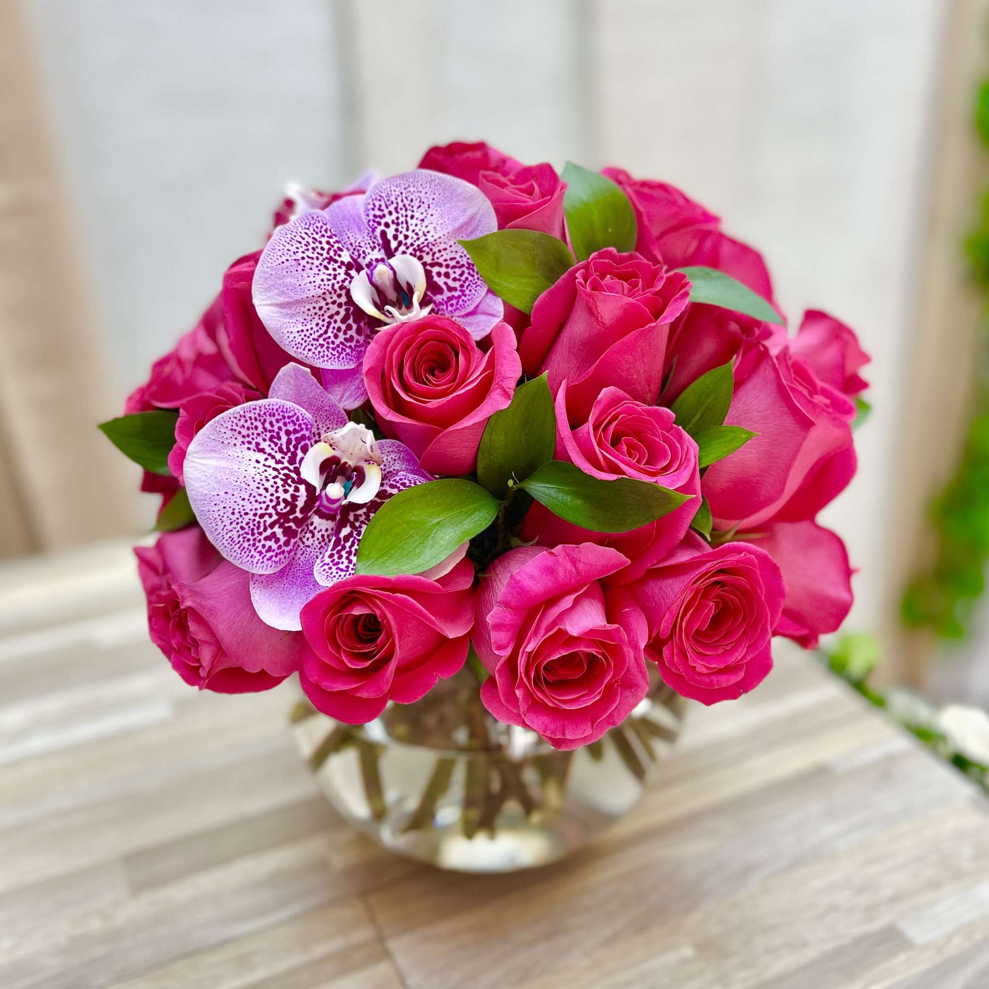 Round arrangement of hot pink roses with speckled lavender orchids in a clear glass bowl vase