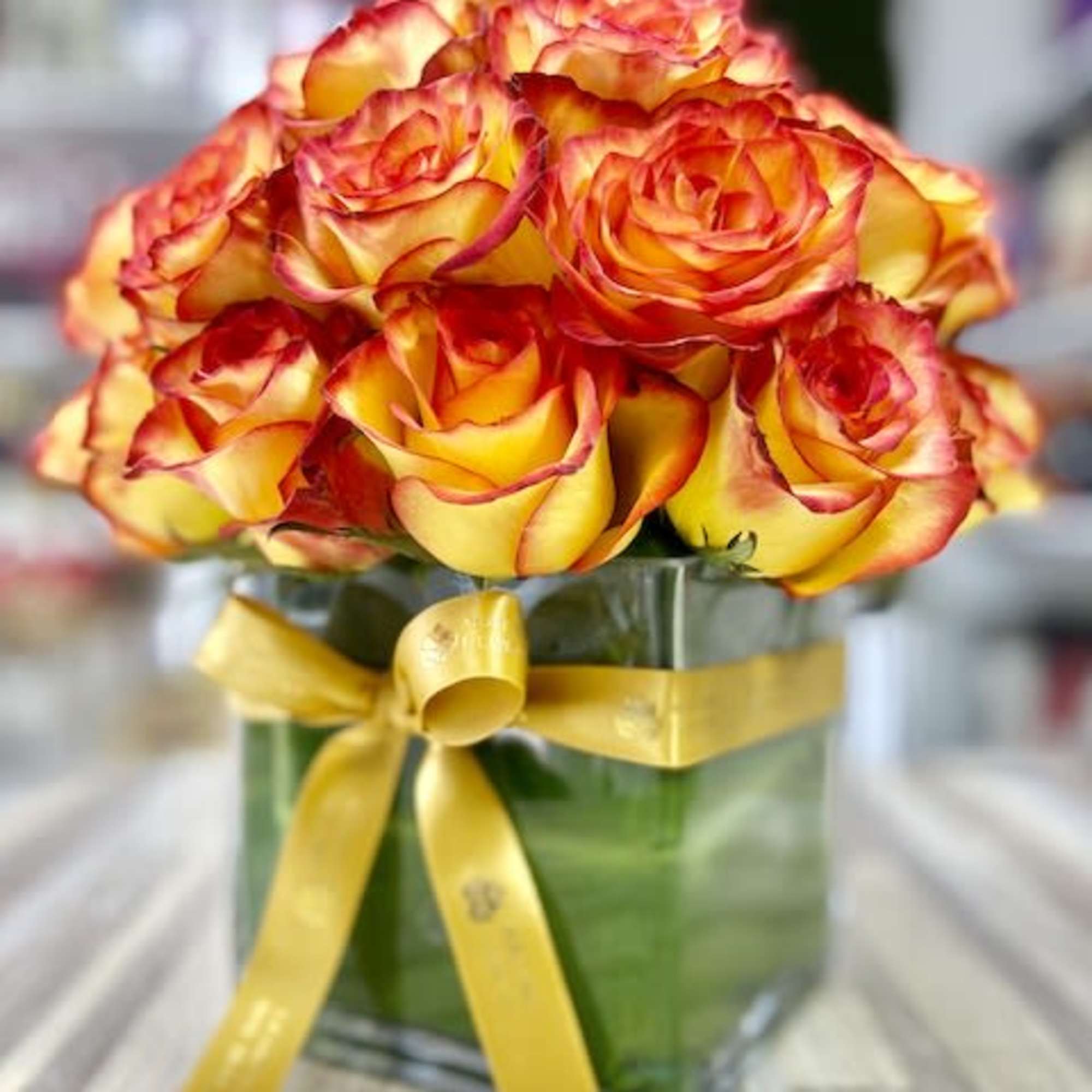 Compact bouquet of yellow roses with red edges in a square glass vase tied with a yellow ribbon