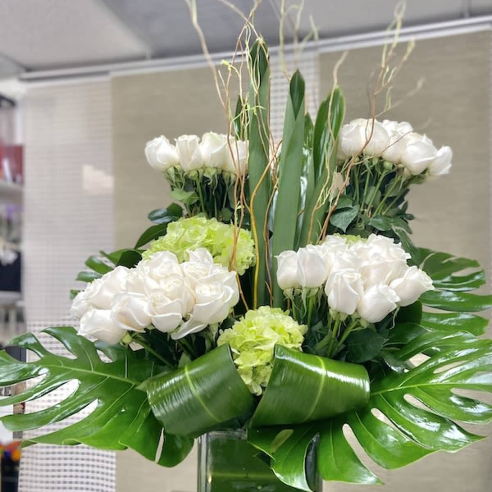 Tall modern arrangement of white roses and green hydrangeas in a square glass vase with large tropical leaves.
