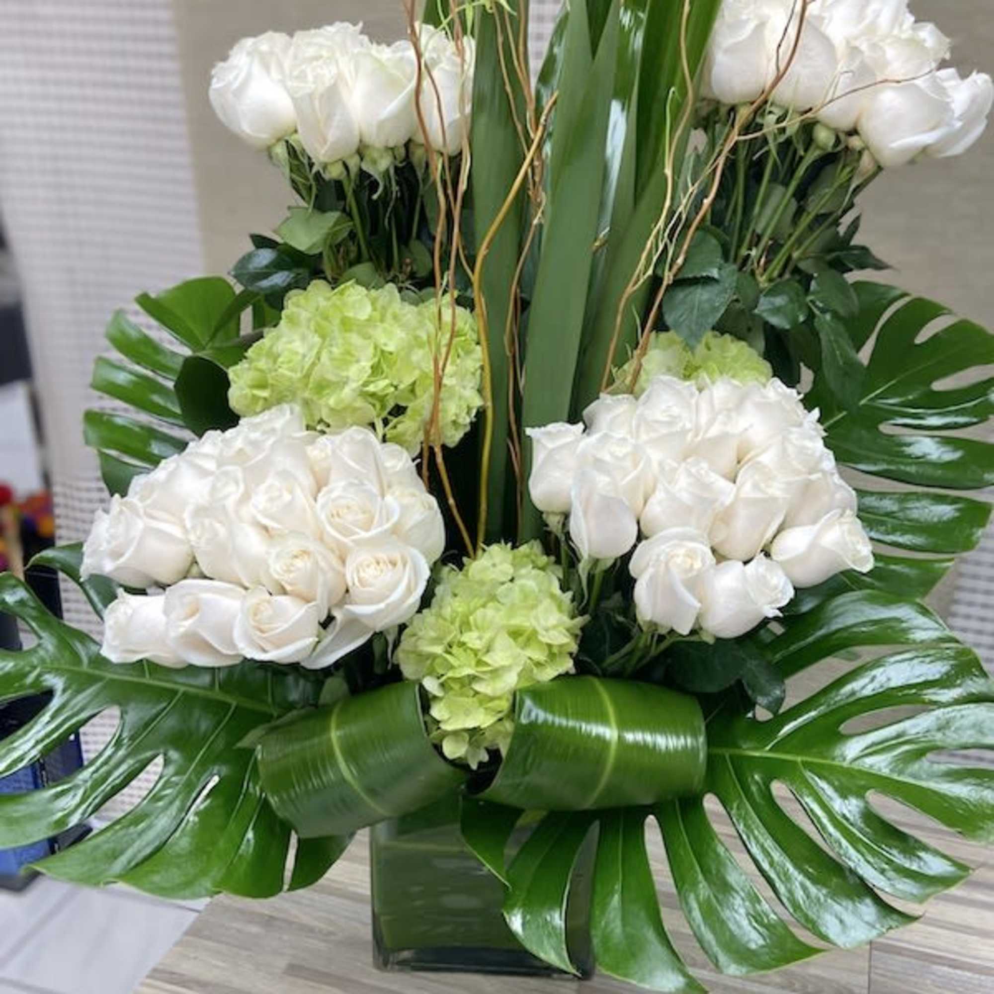 Tall modern arrangement of white roses and green hydrangeas with large tropical leaves in a glass vase