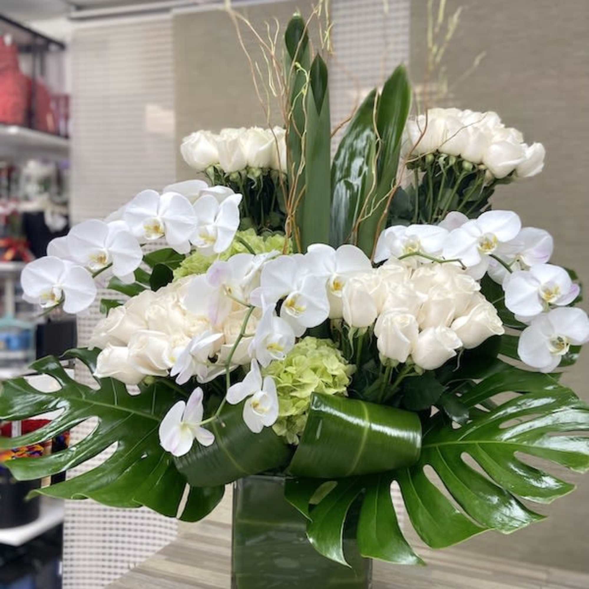 Tall modern arrangement of white roses and orchids with large tropical leaves in a square glass vase
