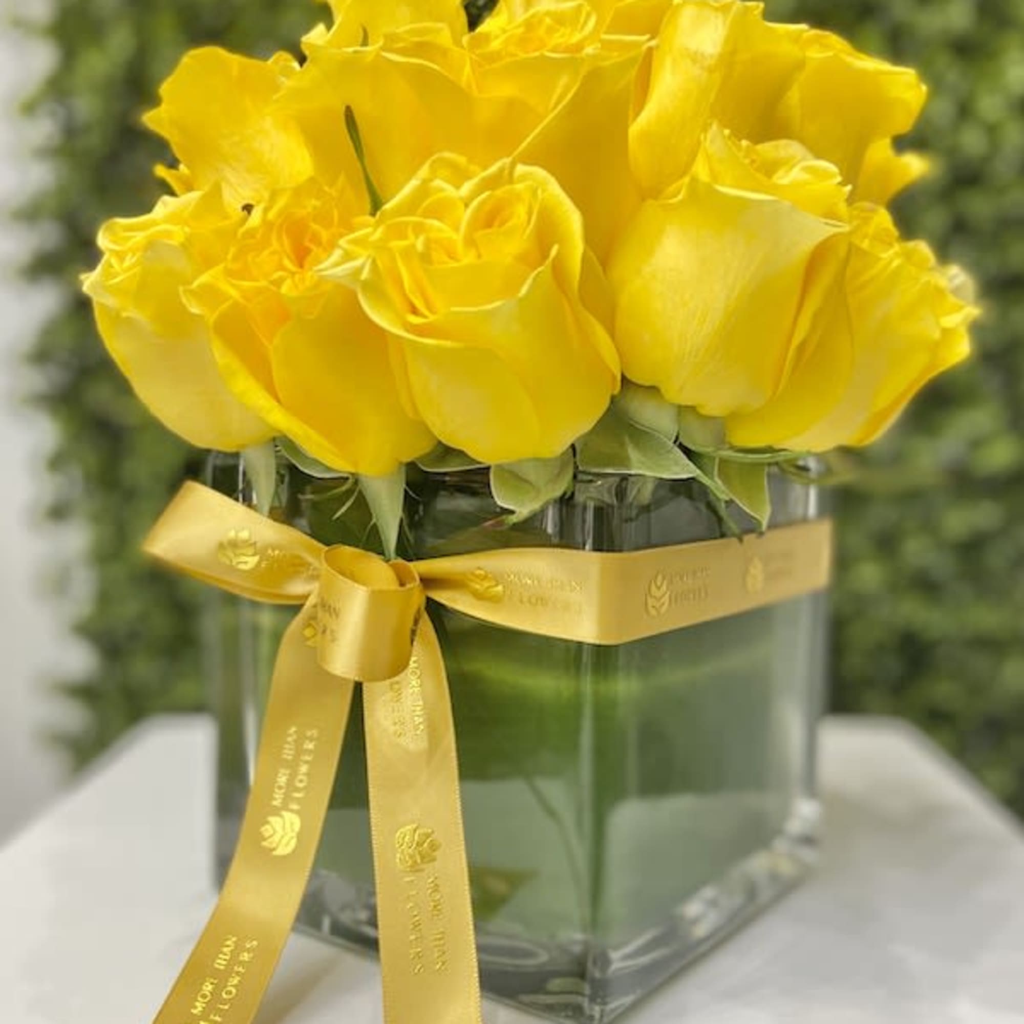 Compact arrangement of bright yellow roses in a clear cube vase with a yellow ribbon bow
