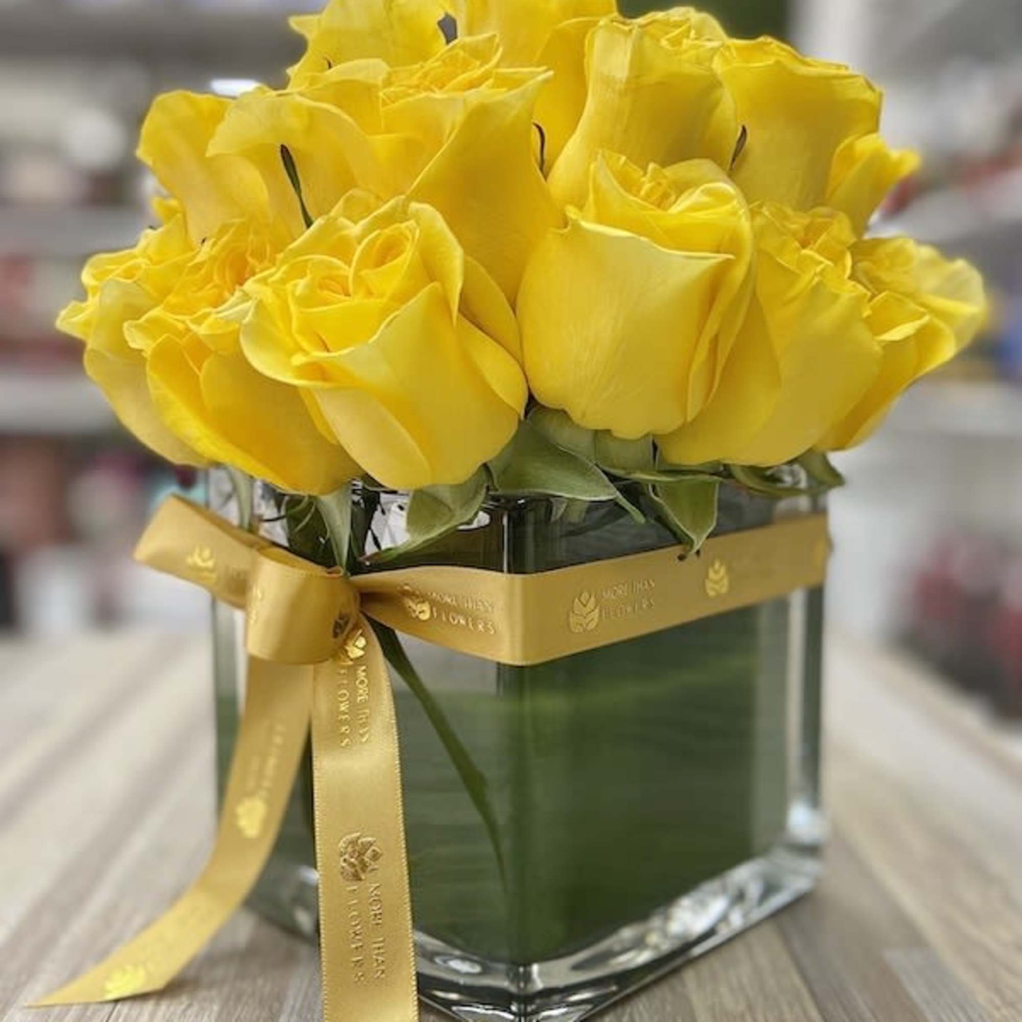 Compact arrangement of yellow roses in a square glass vase wrapped with a yellow ribbon.