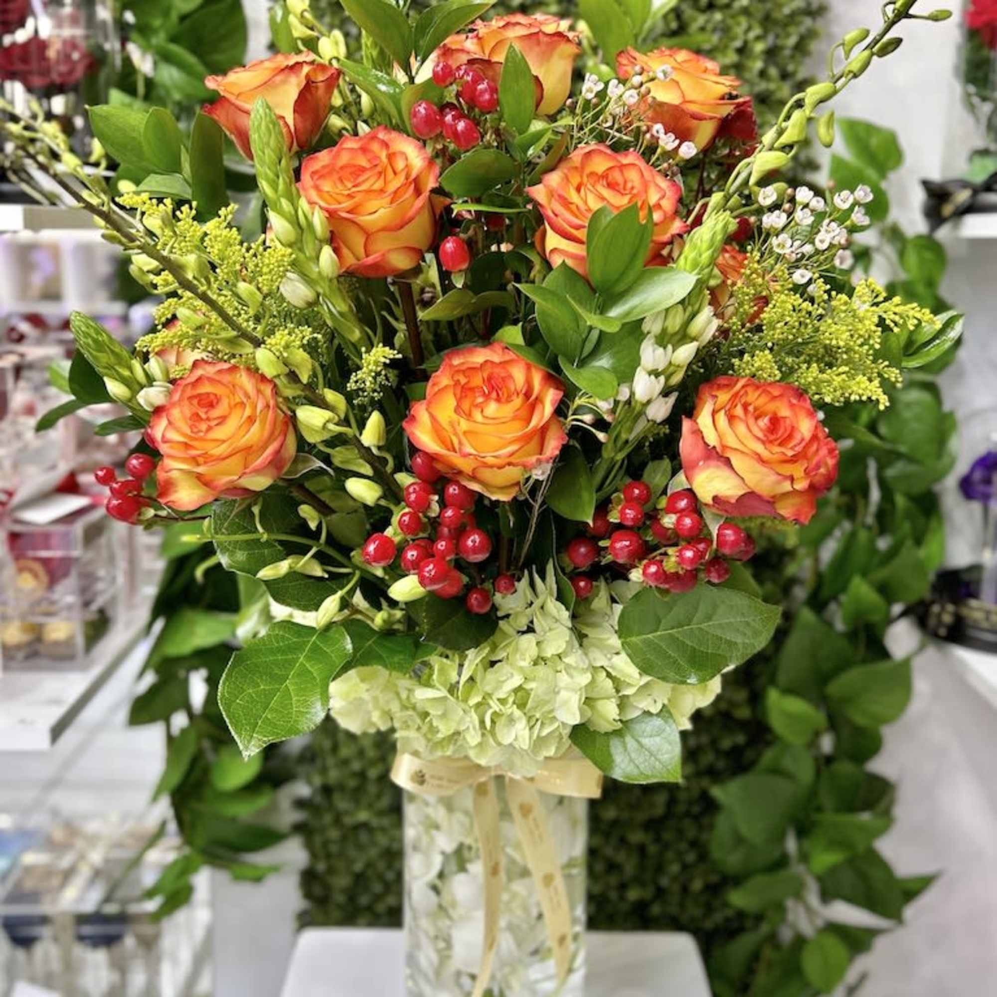 Tall arrangement of orange roses with red berries and green hydrangea in a clear glass vase.