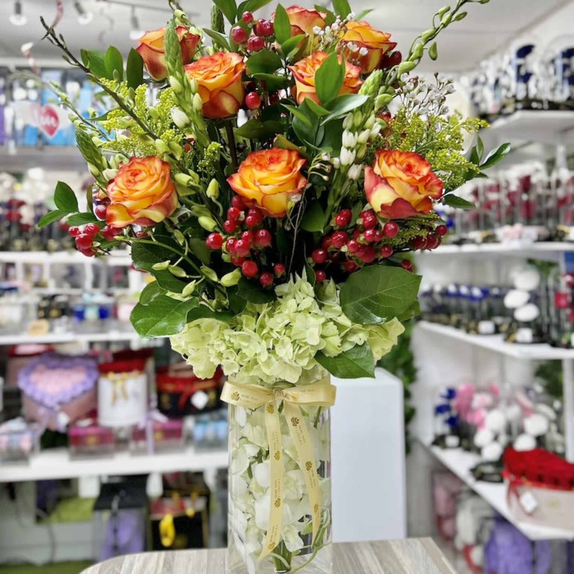Tall arrangement of orange roses, red berries, and green hydrangeas in a clear glass vase with ribbon