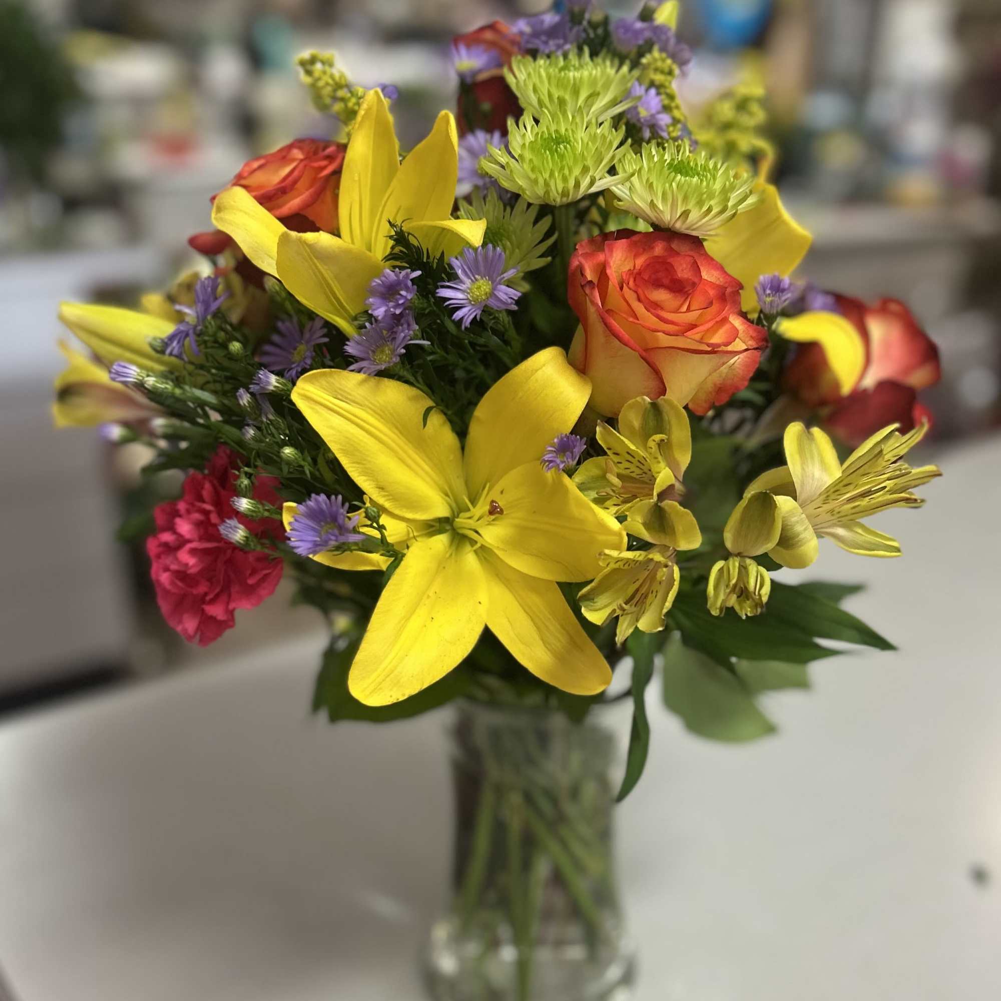 Bouquet of yellow lilies, orange roses, and purple flowers in a glass vase