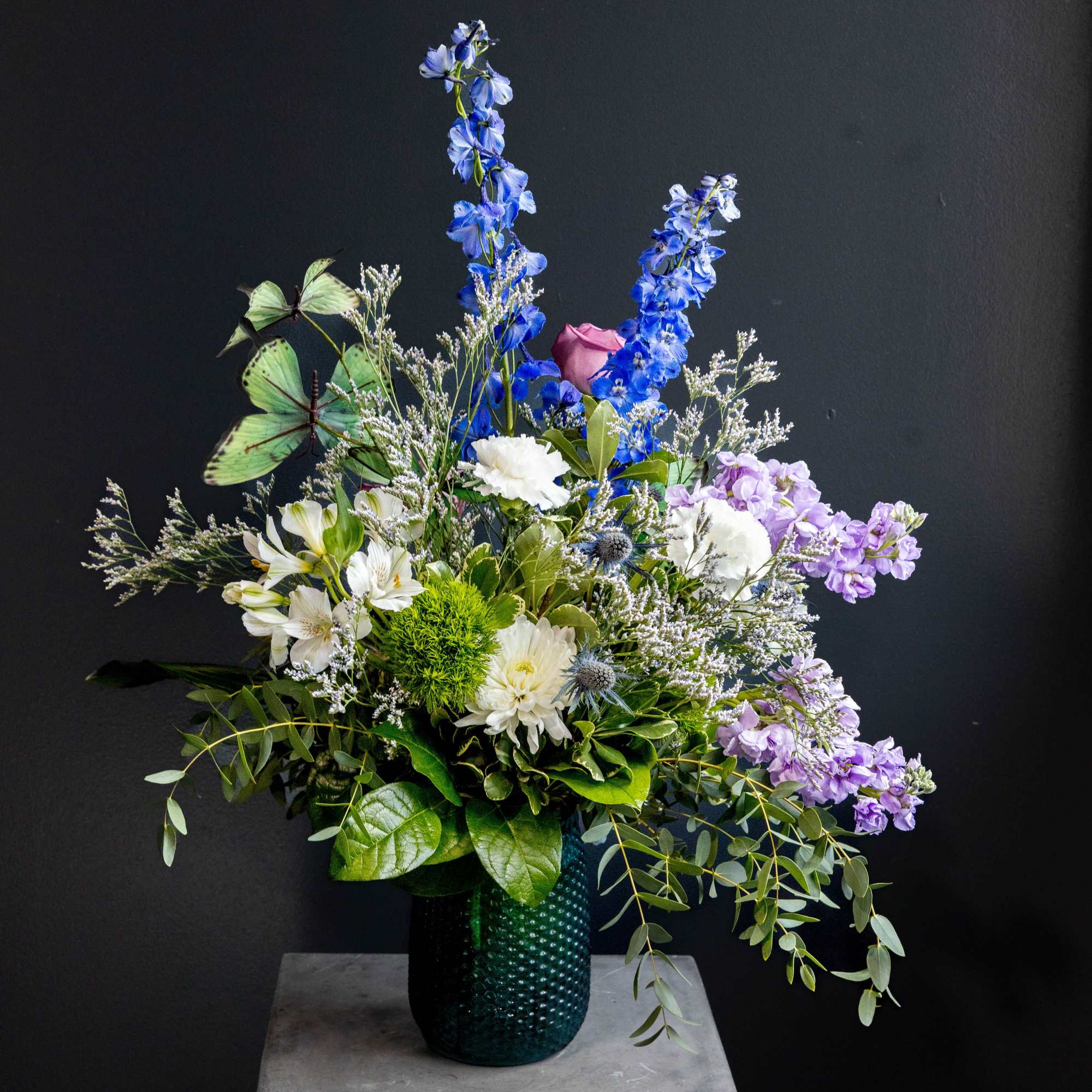 Tall mixed bouquet in a dark green vase with blue, white, and lavender flowers