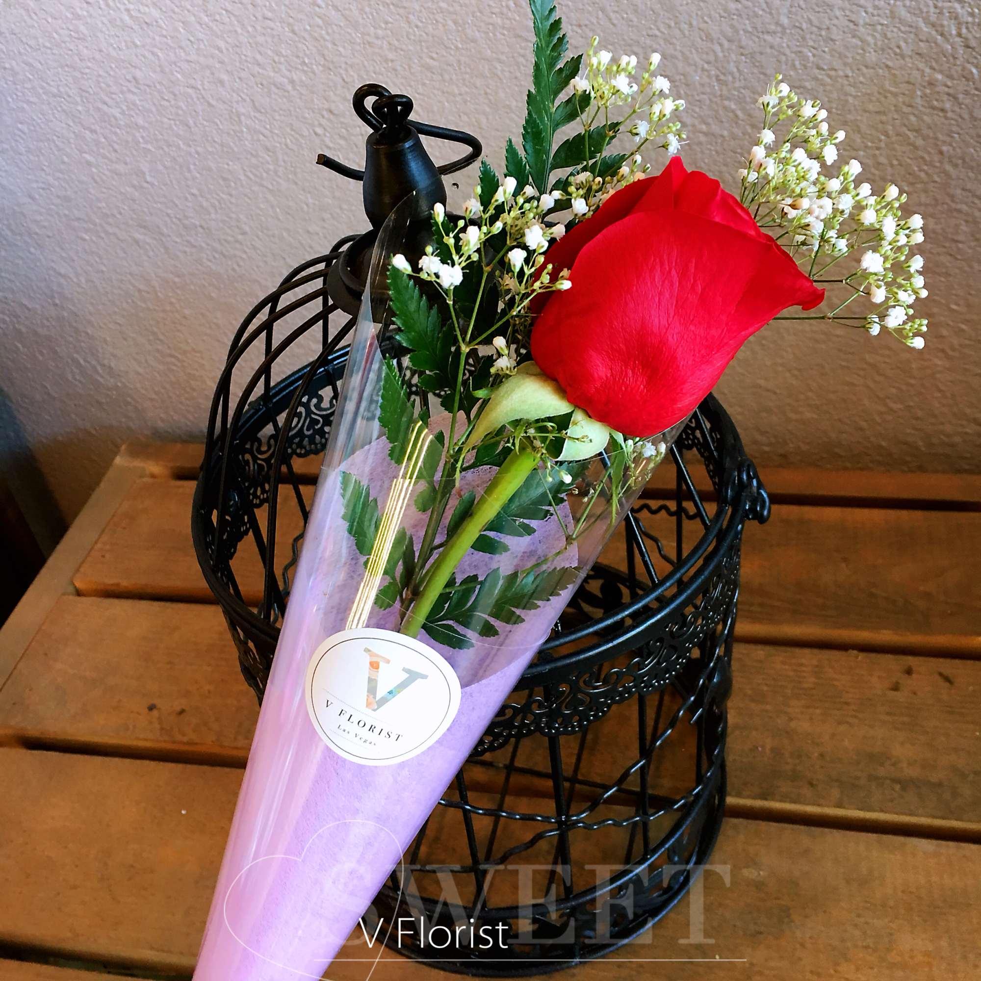 Single red rose with small white blooms wrapped in a purple cone sleeve on a wooden surface.