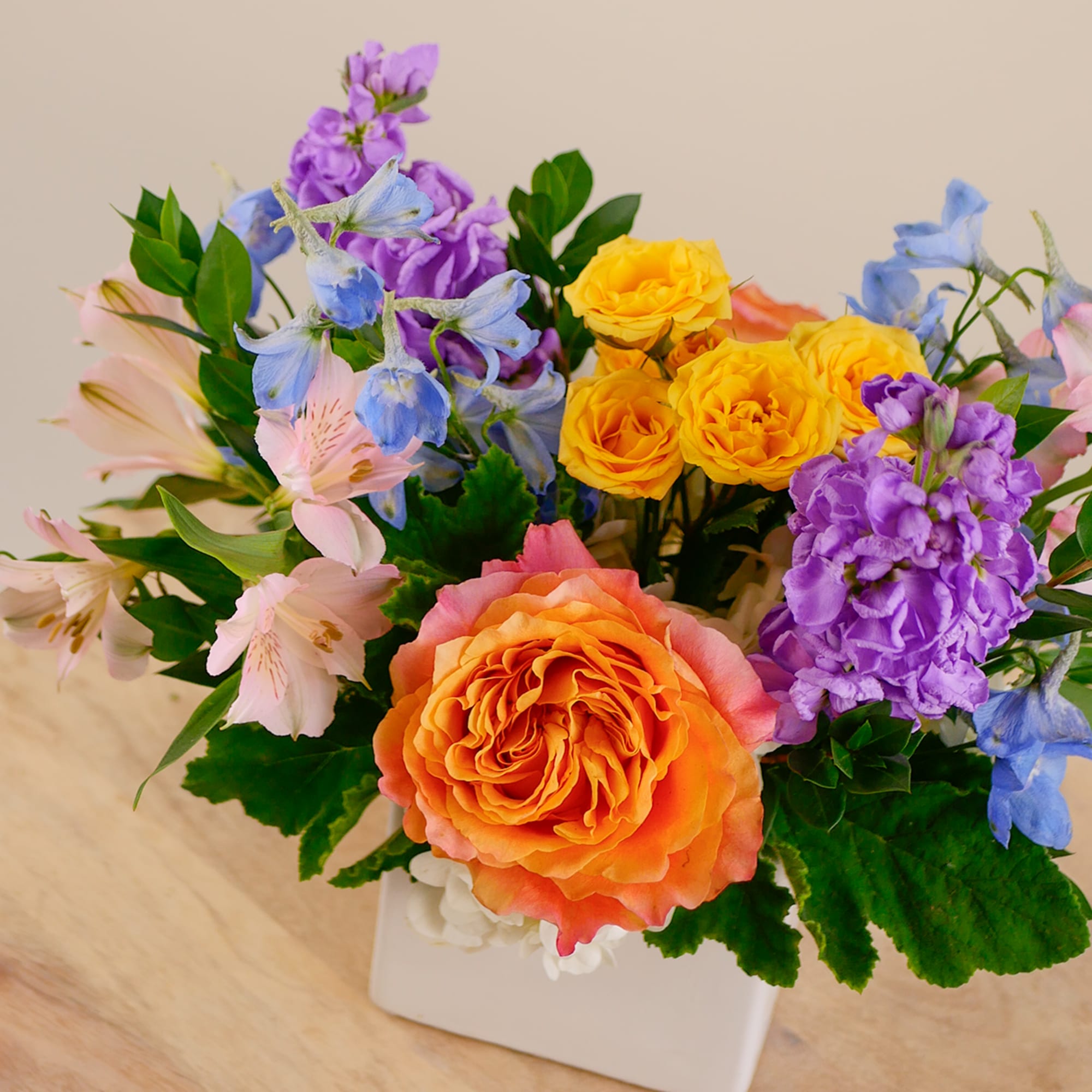 Colorful bouquet of roses, purple blooms, and blue flowers in a white vase