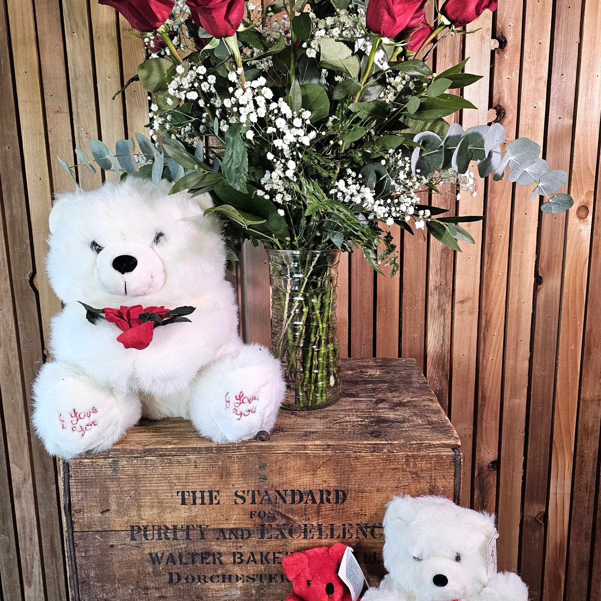 Red roses in a glass vase with two teddy bears on a wooden crate