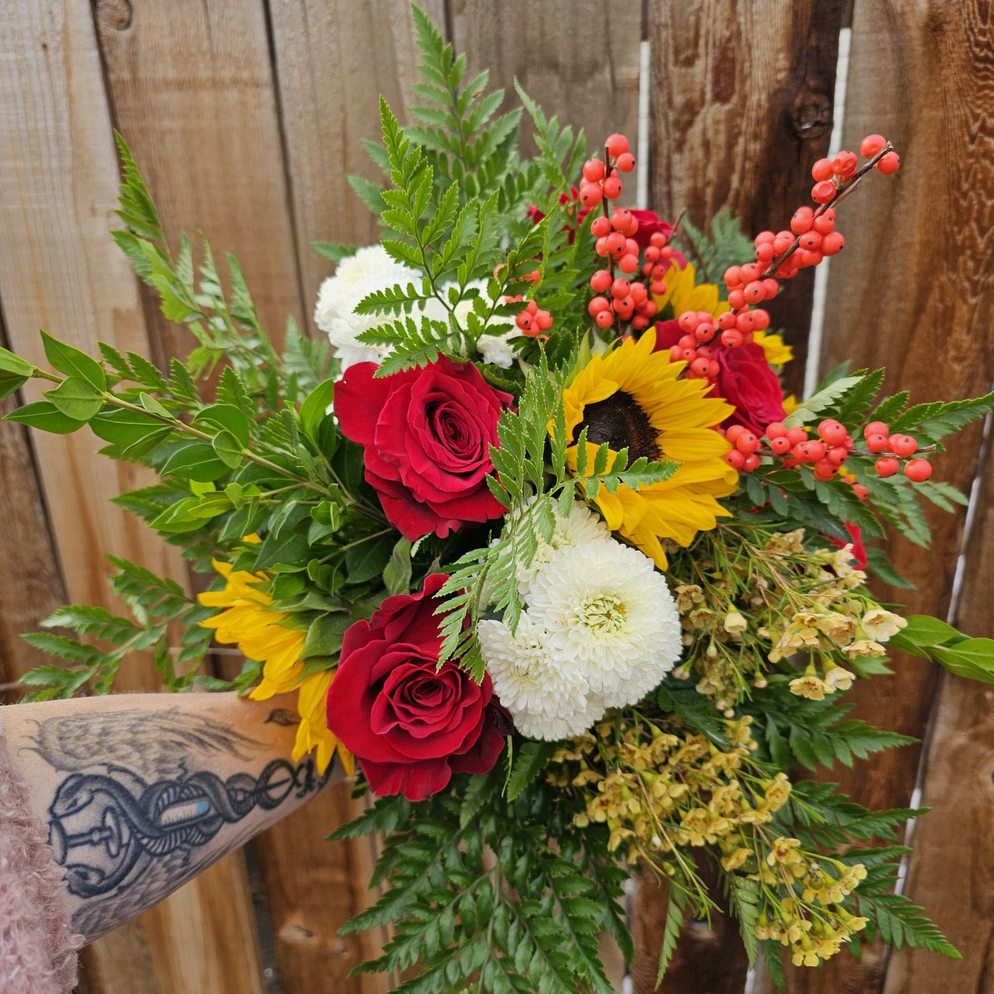 Handheld bouquet of red roses, sunflowers, and white blooms with berry accents