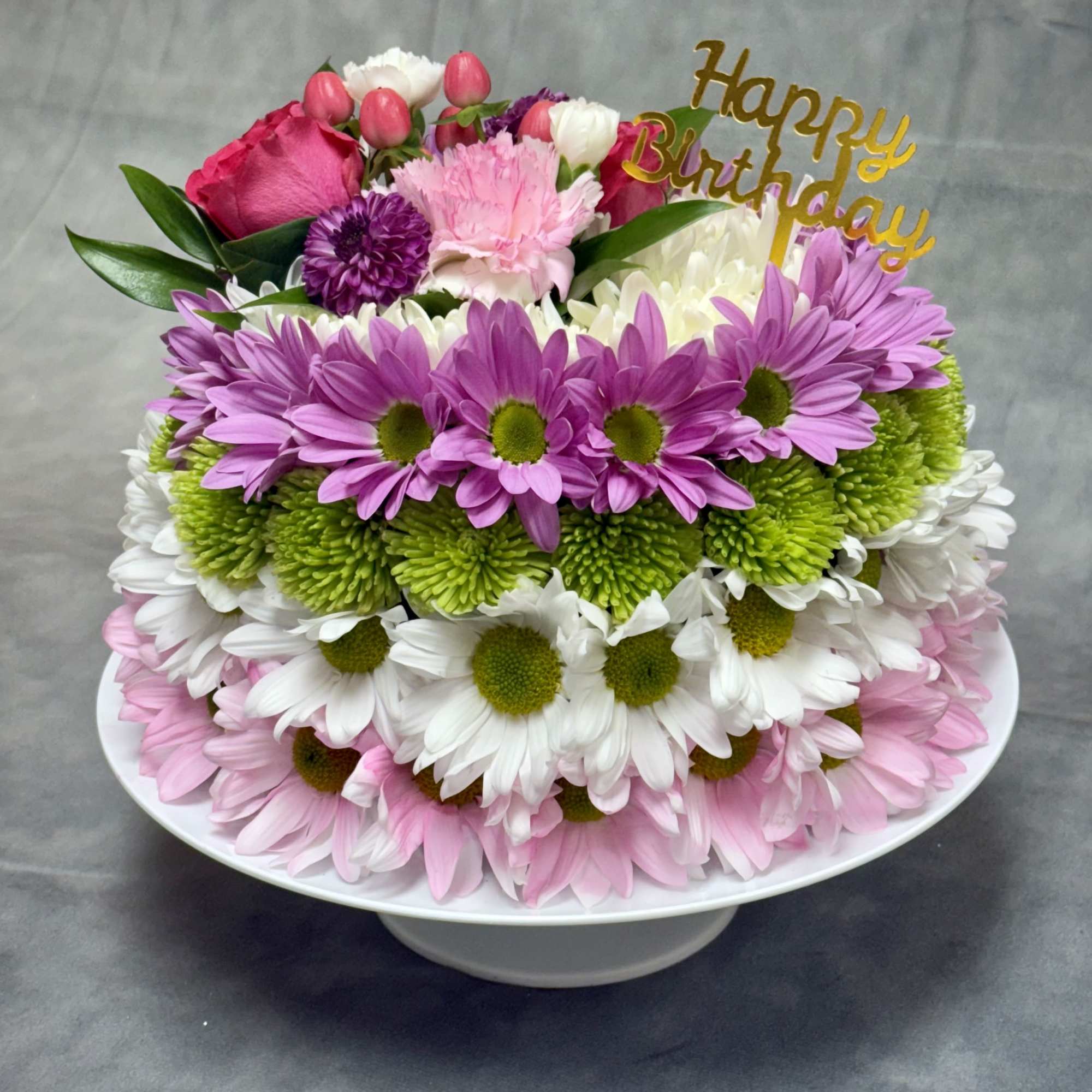 Tiered birthday cake covered with daisies and a gold topper