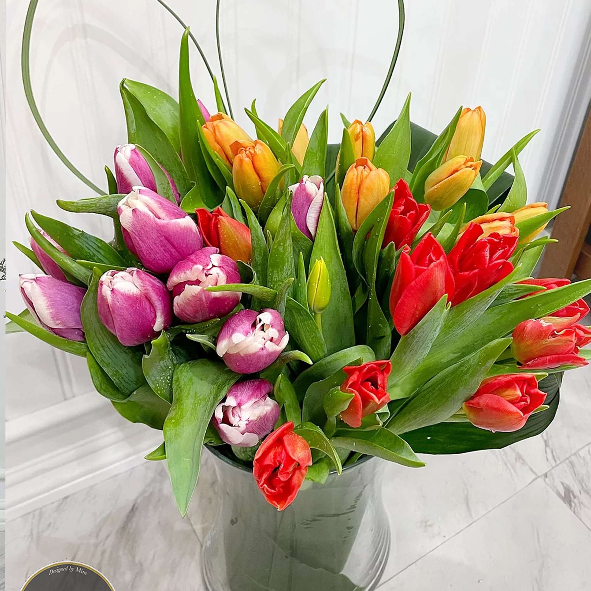 Mixed red, orange, and purple tulips in a clear glass vase with heart-shaped greenery loops